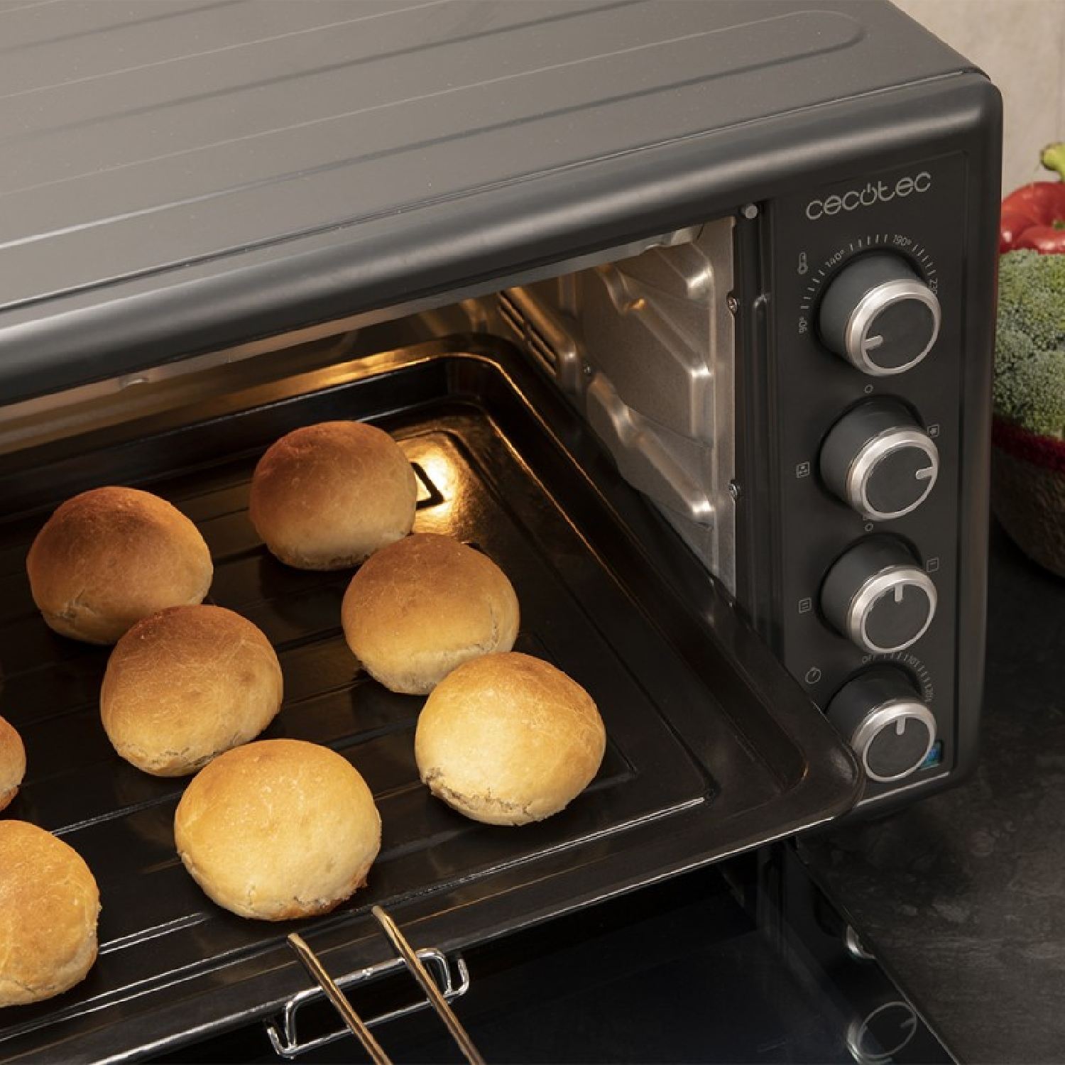 Un horno con bollos horneados dentro. El horno es negro y tiene varias perillas. Una cesta de verduras está en el fondo.