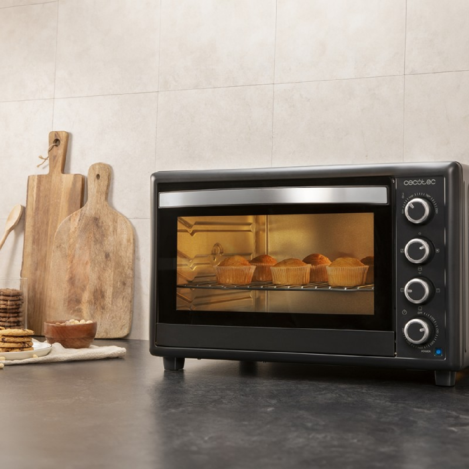 Un horno negro con magdalenas dentro está sobre una encimera. Tablas de madera y galletas están cerca. Fondo de baldosas beige.