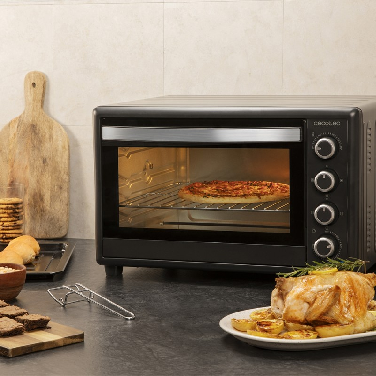 Una pizza se hornea en un horno negro. Un pollo asado y otros alimentos están en la encimera.