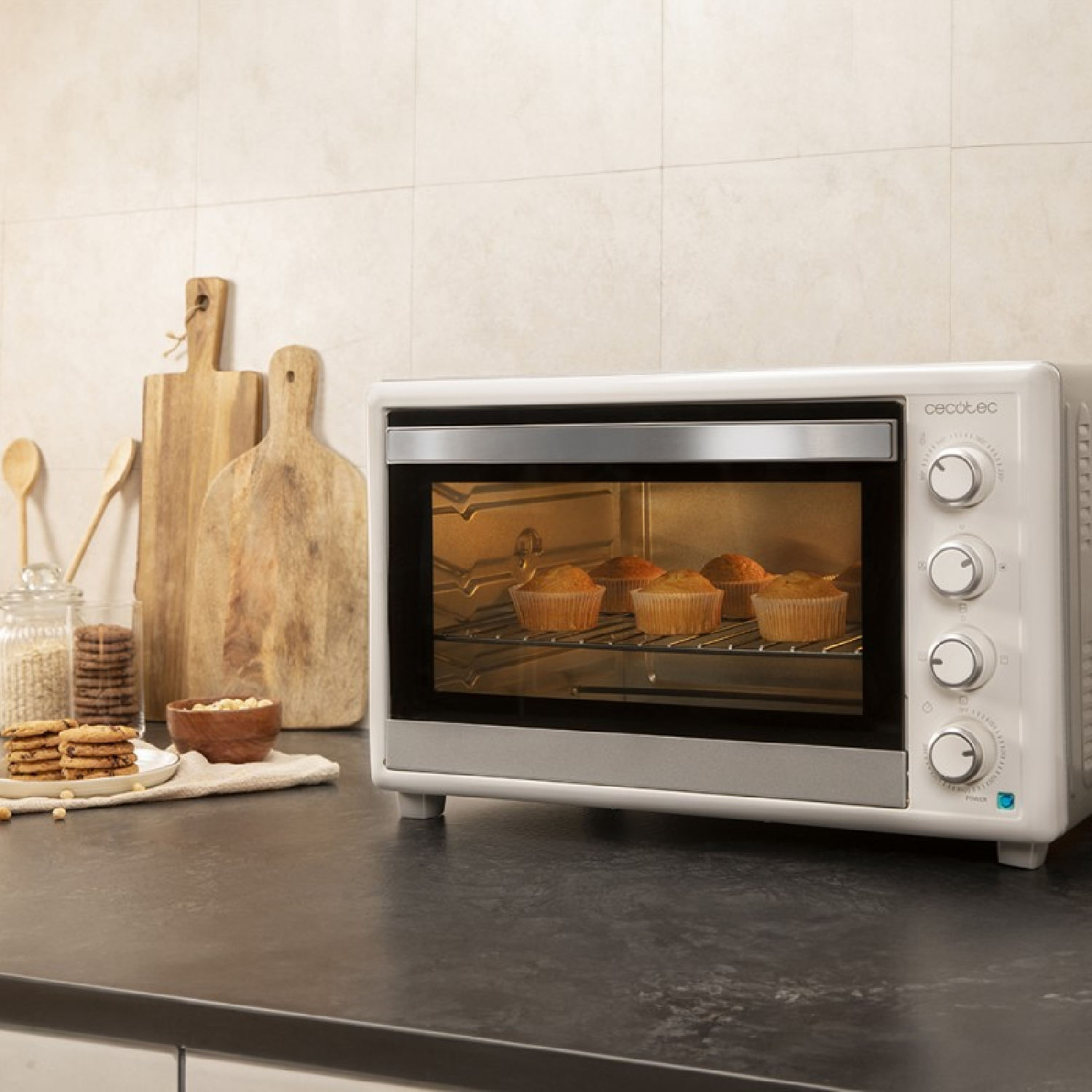 Een witte oven met muffins staat op een zwart aanrecht. Houten planken en koekjes staan ook op het aanrecht.