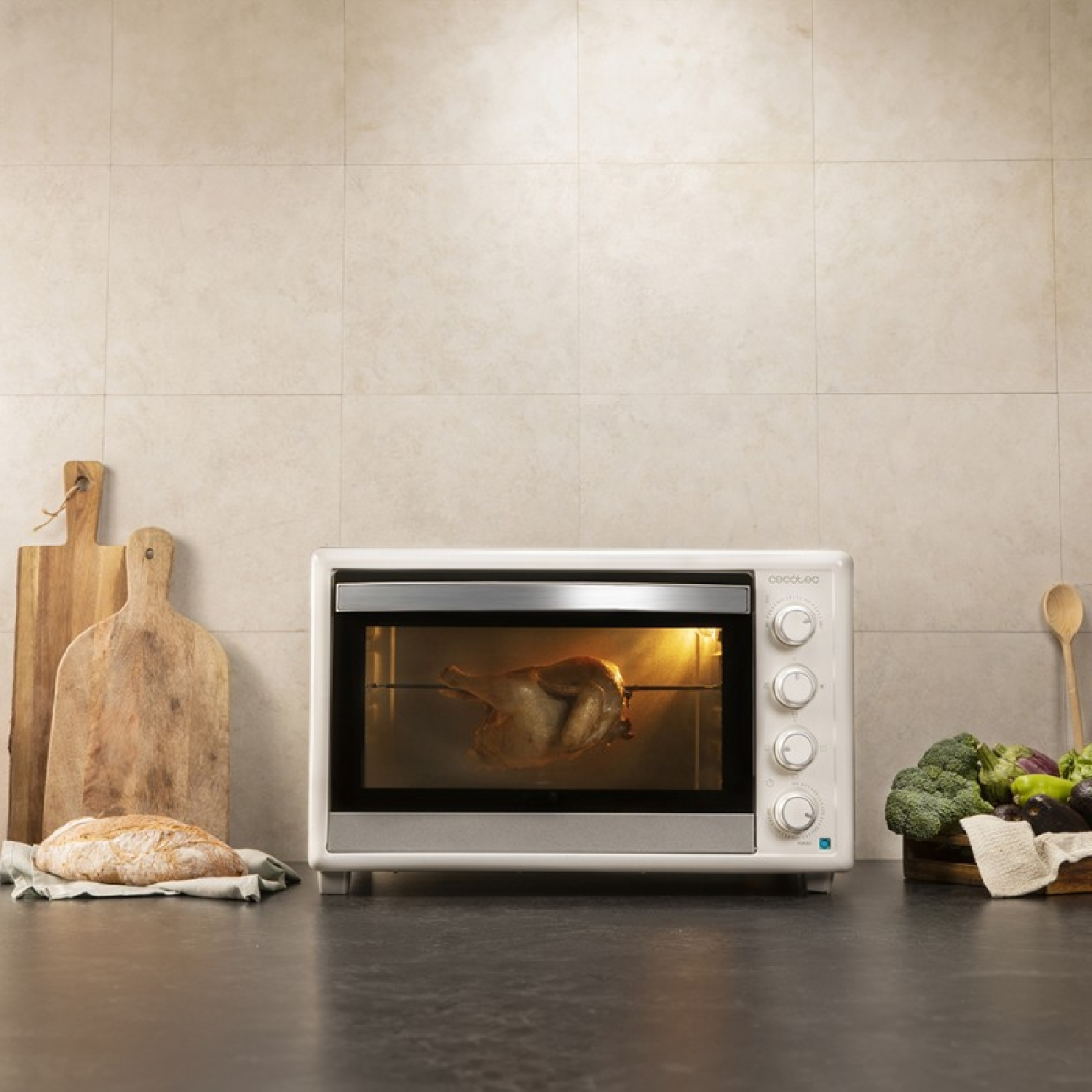 Een witte oven met een gebraden kip erin. Brood en snijplanken links. Groenten rechts.