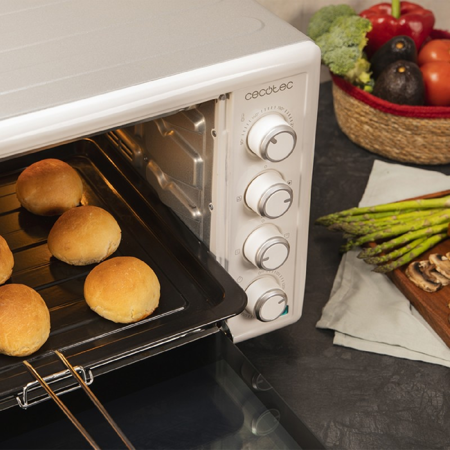 Een oven met gebakken broodjes erin. Groenten zitten in een mand ernaast. Asperges en champignons op een plank.