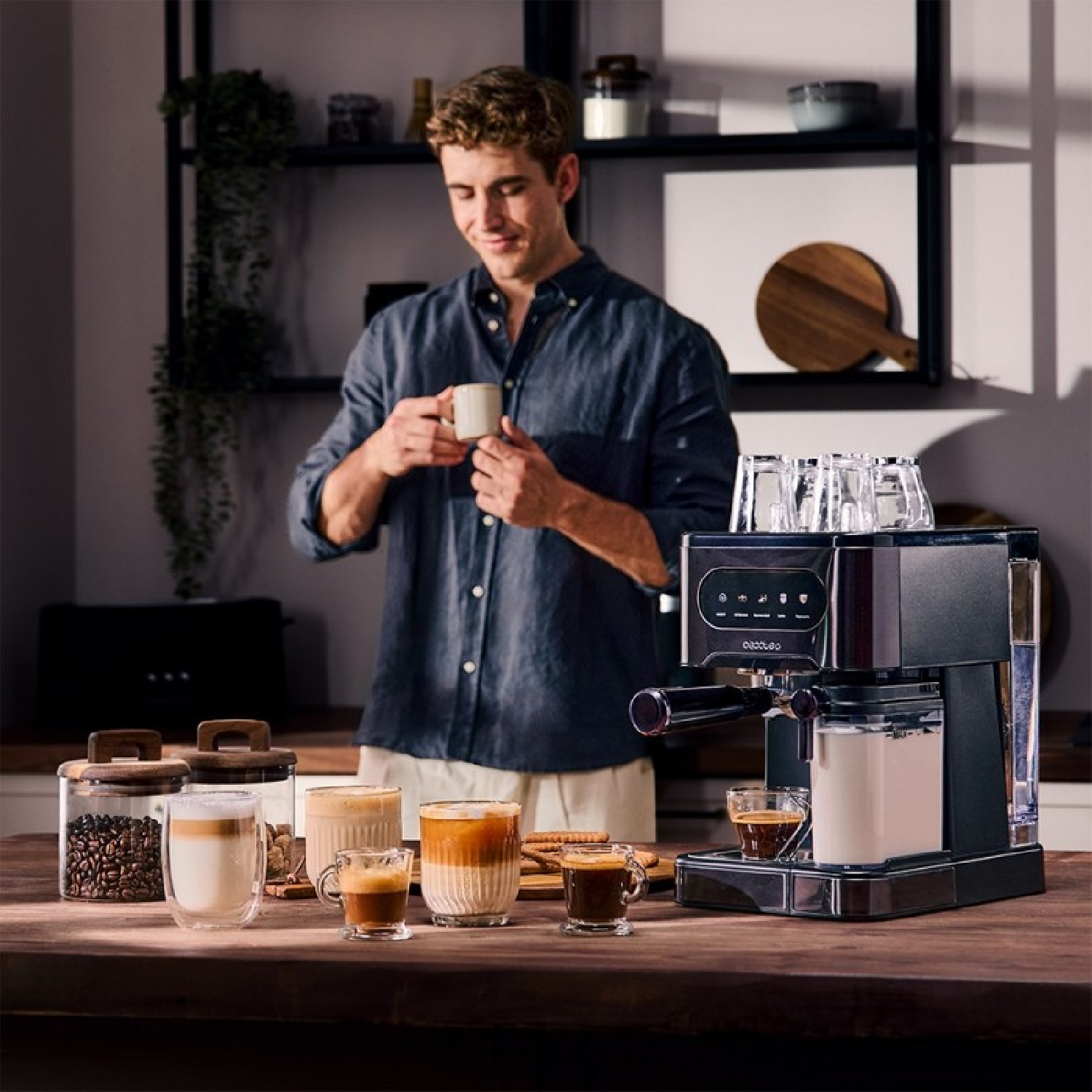 Un hombre sostiene una taza cerca de una máquina de espresso. Tazas de café e ingredientes en una encimera de madera, con estantes detrás.