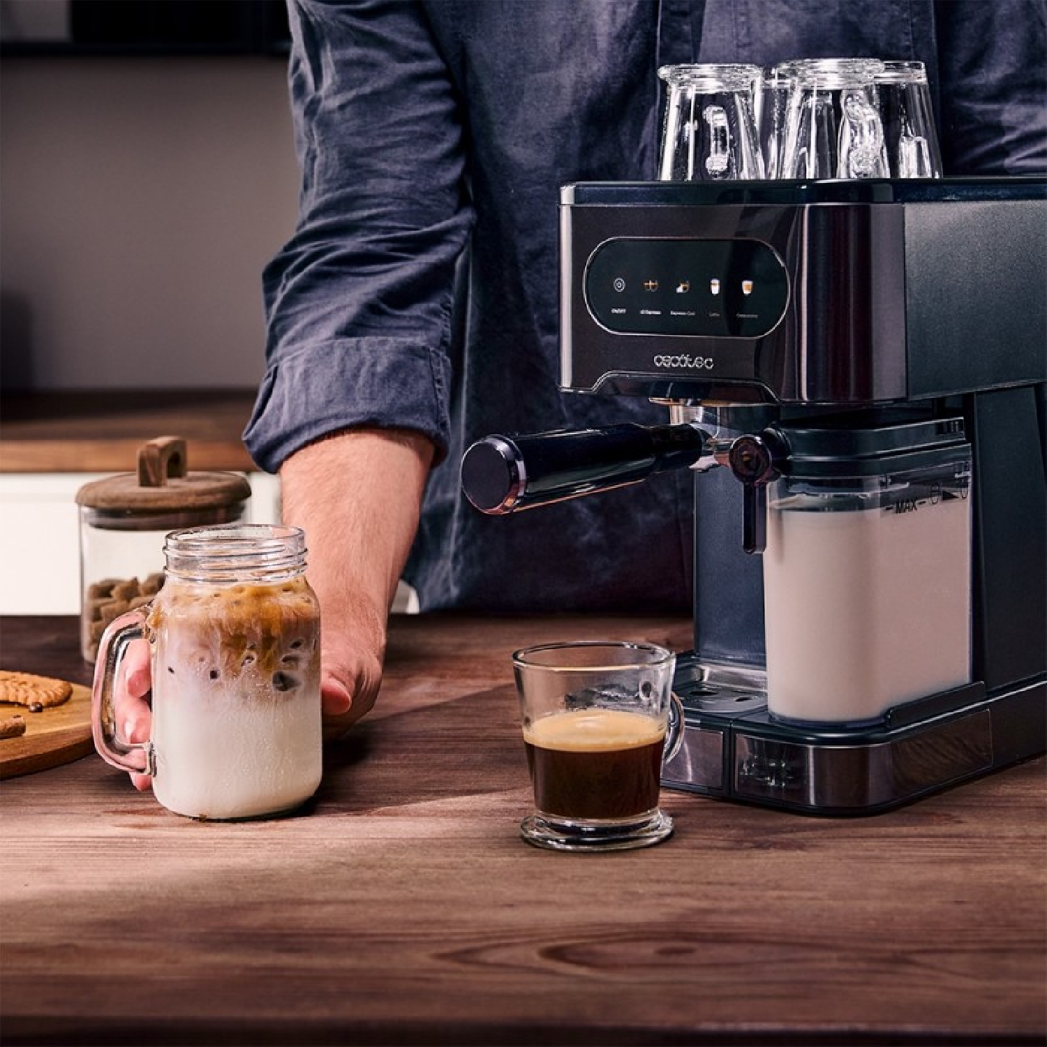 Una persona prepara café con una máquina. Hay un vaso de café, un vaso con leche y café y un tarro con galletas.