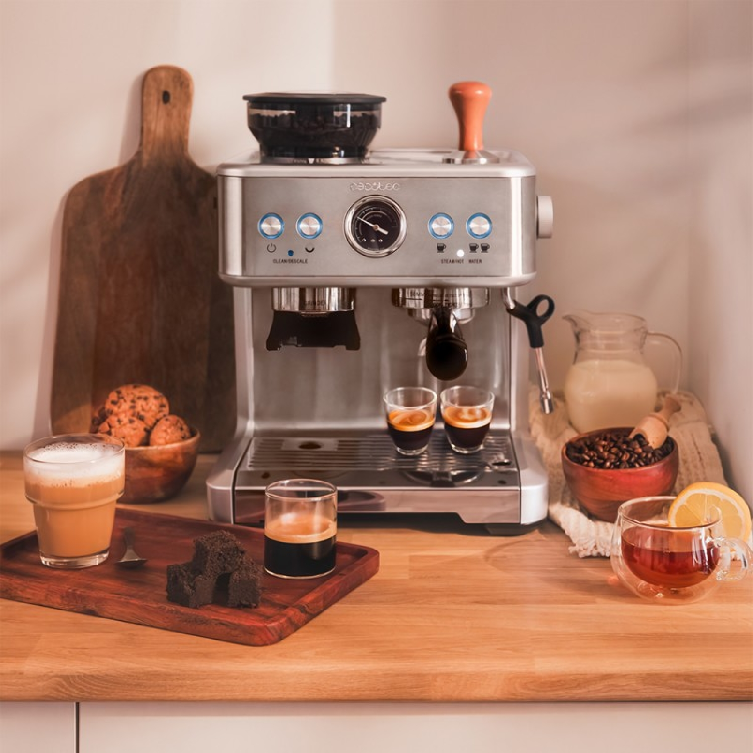 Cafetera con café espresso, té, galletas y leche. Hay una tabla de madera y una tabla de cortar, todo sobre una superficie de madera.