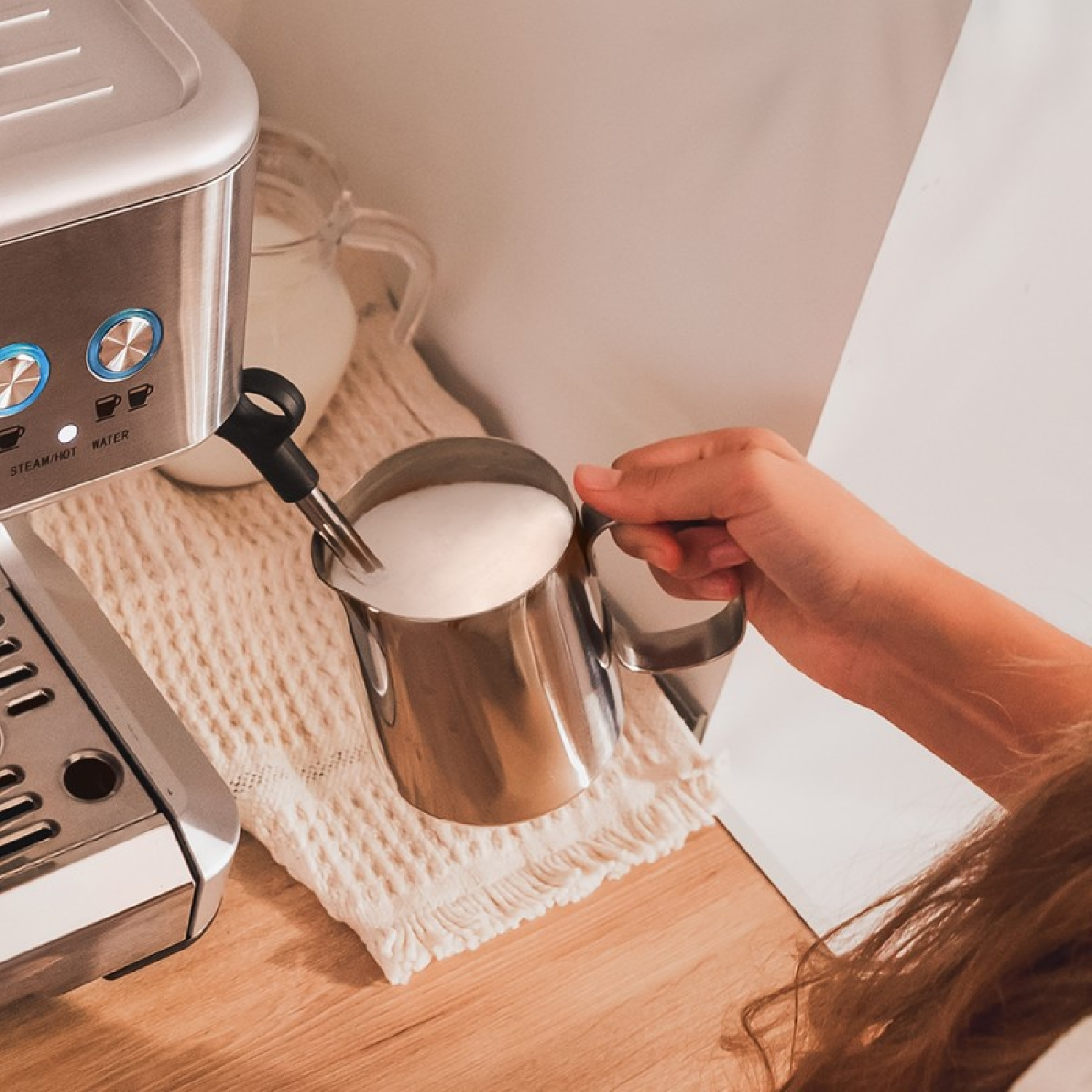 Ręka spienia mleko w metalowym dzbanku. Po lewej ekspres do kawy. W tle dzbanek z mlekiem.