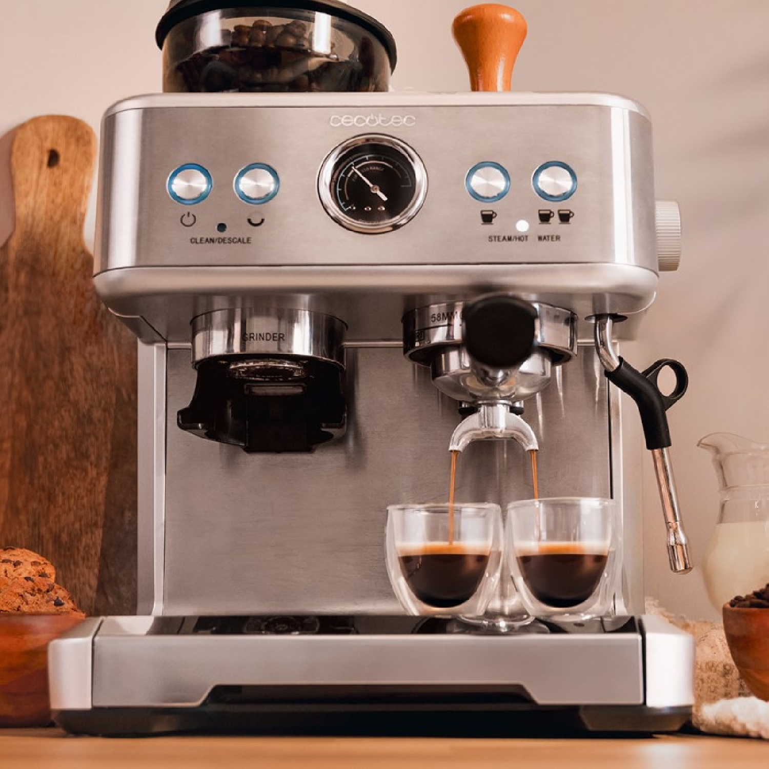 Máquina de espresso dispensando café en dos vasos de vidrio transparente. Se ve una tabla de cortar de madera y cuencos con galletas y frijoles.