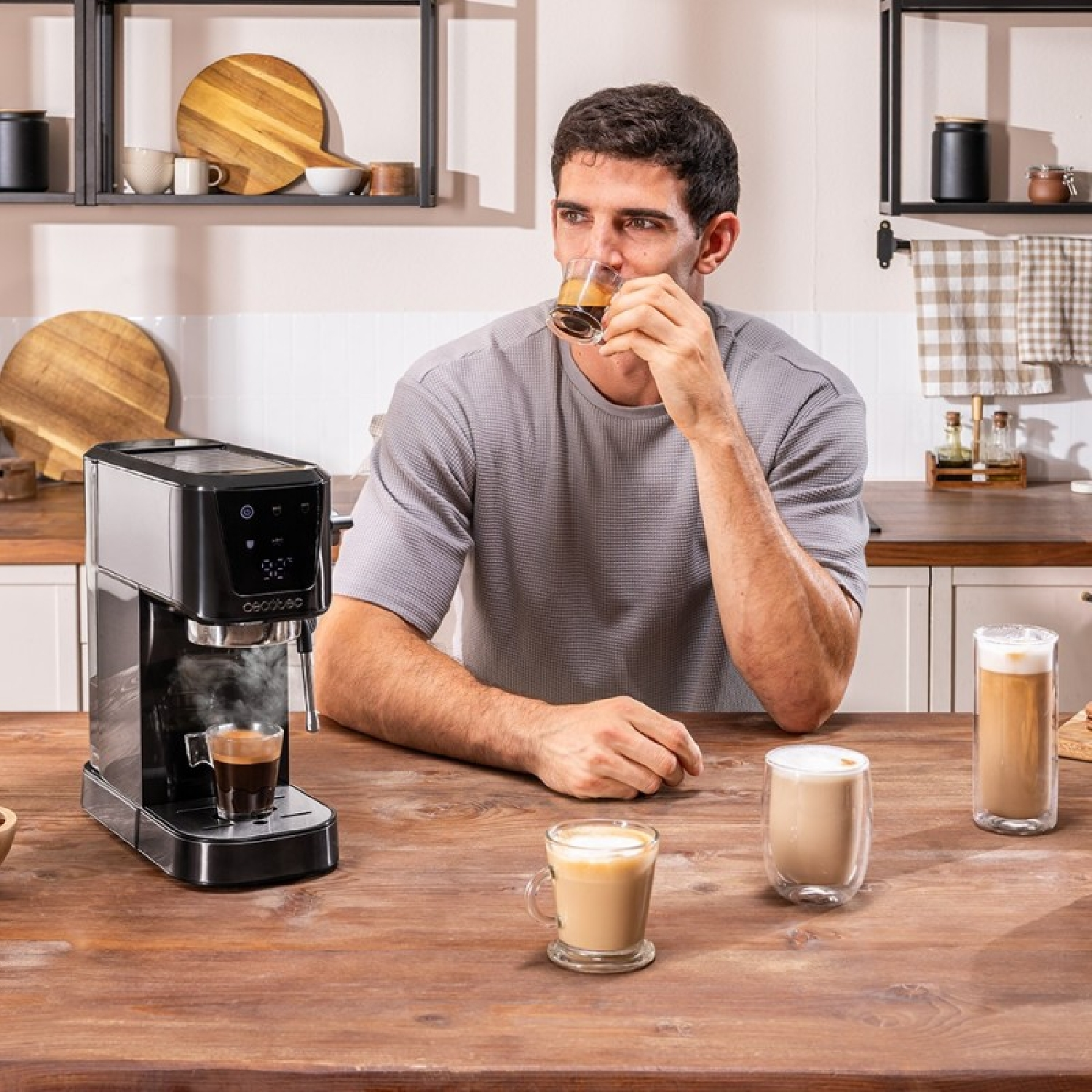 Un hombre bebe espresso. Máquina de espresso a la izquierda, varias bebidas de café en la mesa. Fondo de cocina con estantes.