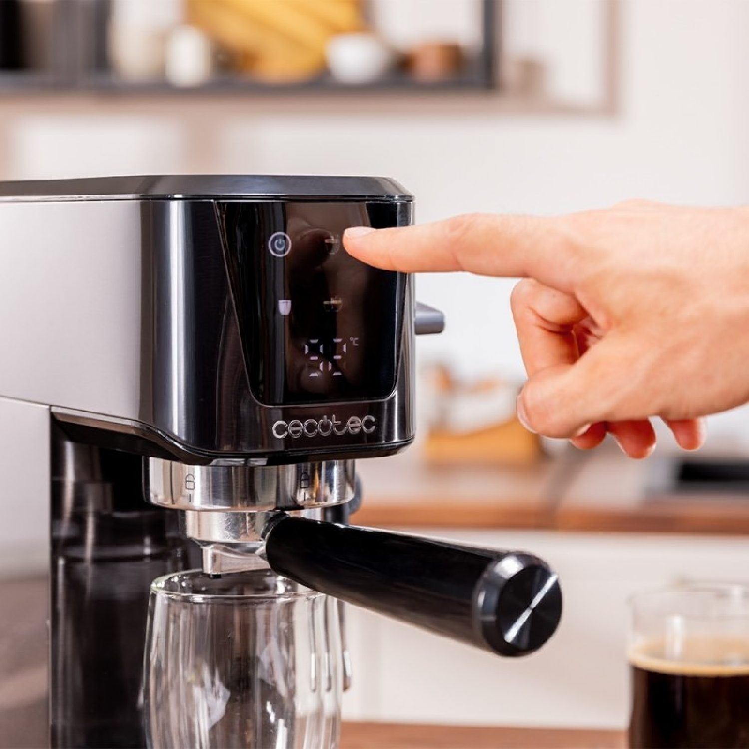 Una mano presionando un botón en una cafetera con un vaso debajo. Una taza de café está en la mesa. La máquina es negra y plateada.