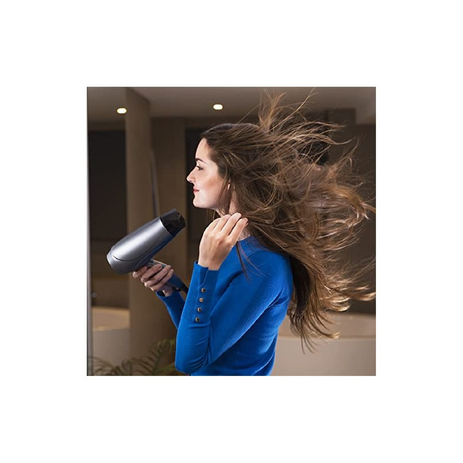 Una donna con un top blu asciuga i suoi lunghi capelli castani con un asciugacapelli argentato. I suoi capelli volano.