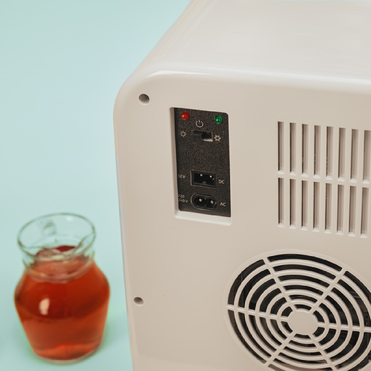 Un mini-refrigerador blanco con una jarra de vidrio con líquido rojo al frente. El refrigerador tiene un panel de alimentación y un ventilador. El fondo es azul claro.
