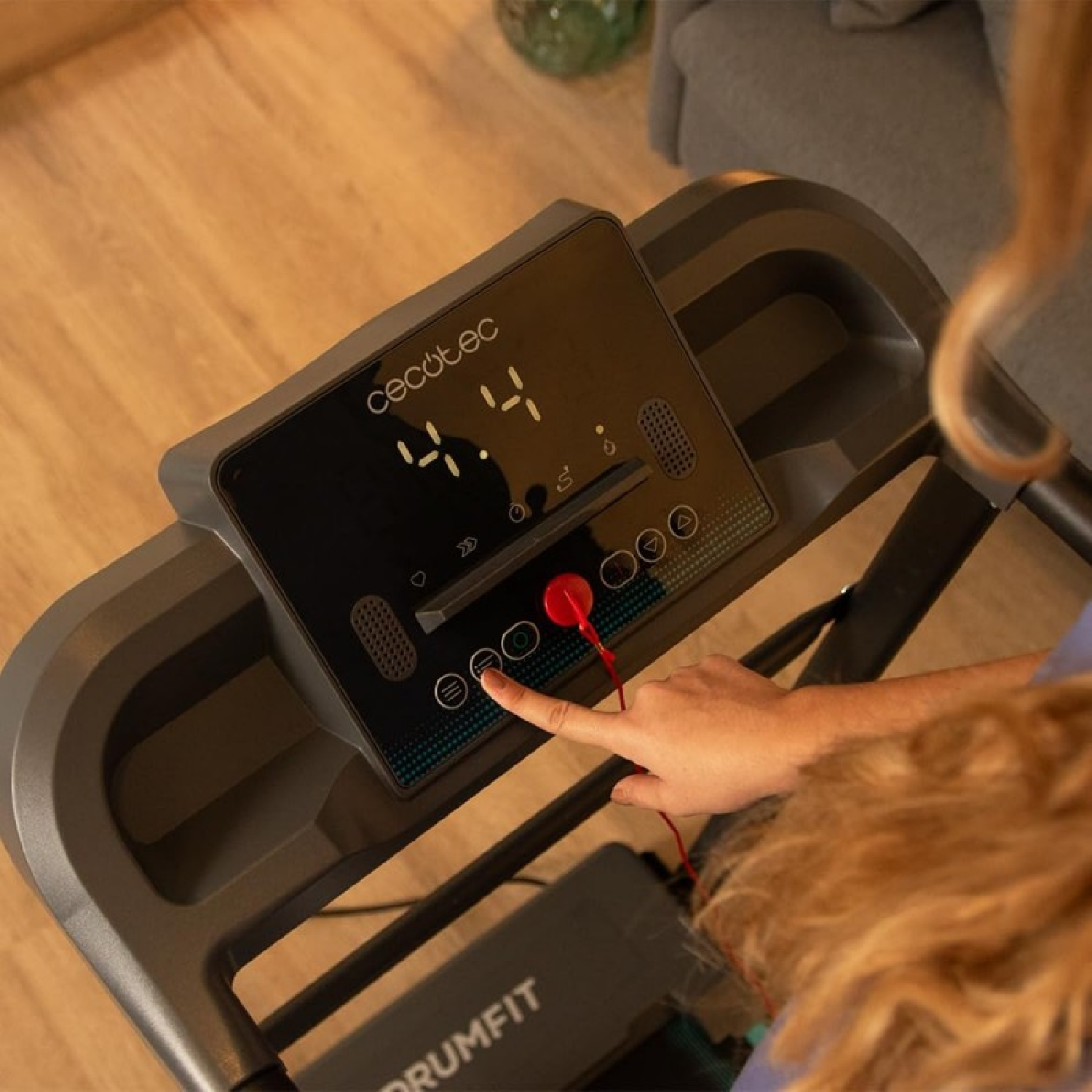 Palec naciska przycisk na konsoli bieżni. Wyświetlacz pokazuje '4.4'. Szara bieżnia z czerwonym kluczem bezpieczeństwa.