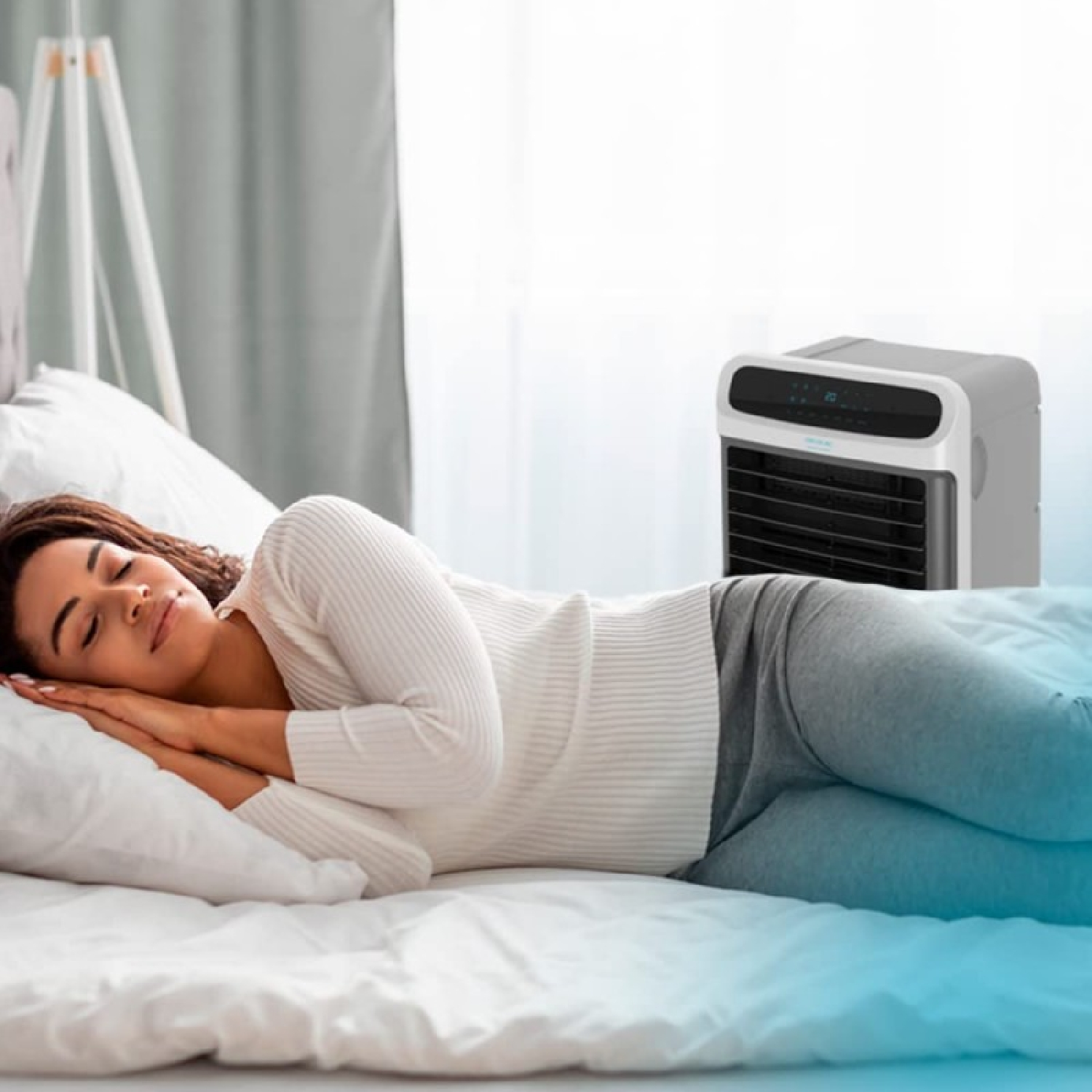 Een vrouw slaapt in bed. Een witte luchtkoeler staat naast het bed. De vrouw draagt een wit shirt en een grijze broek.