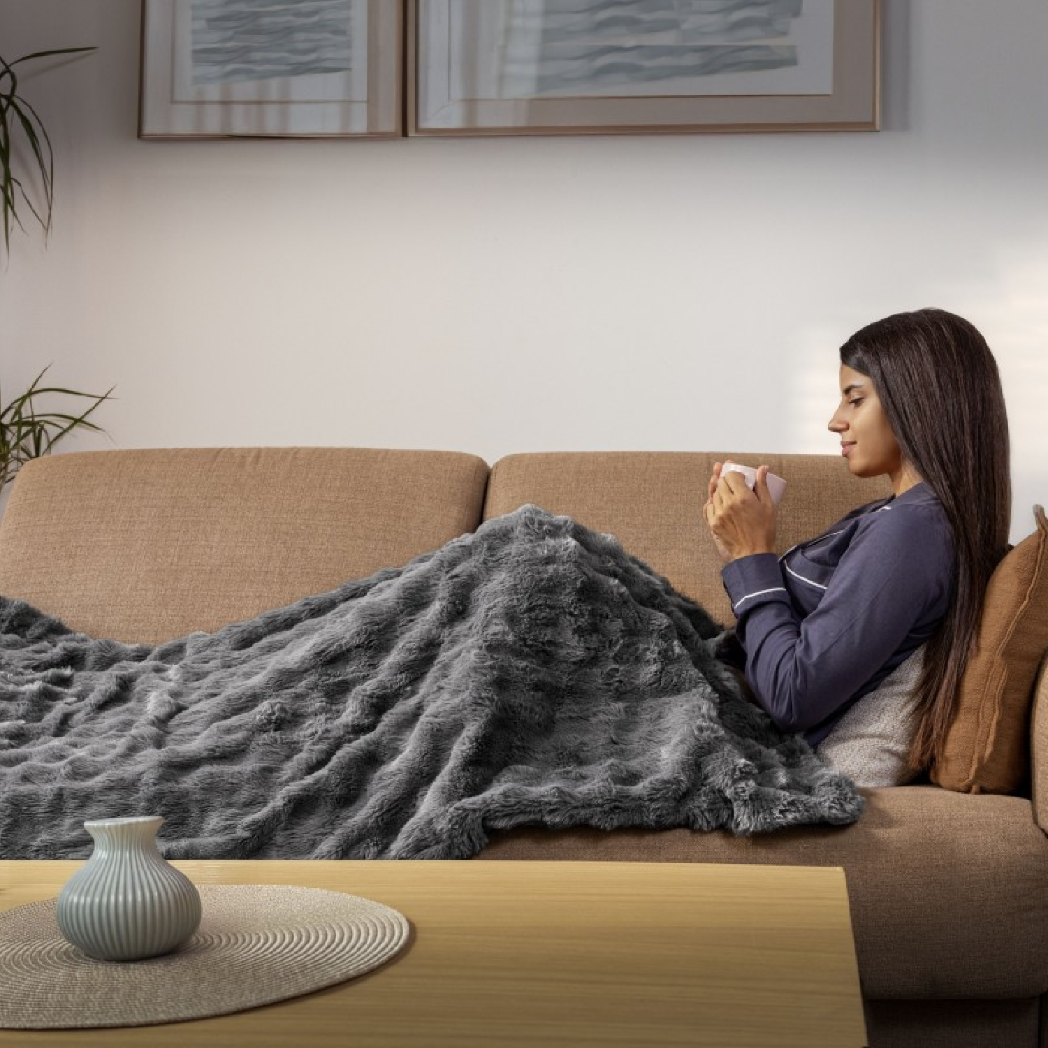 Una mujer en pijama está sentada en un sofá, cubierta con una manta, sosteniendo una taza rosa. Un jarrón está sobre una mesa delante.