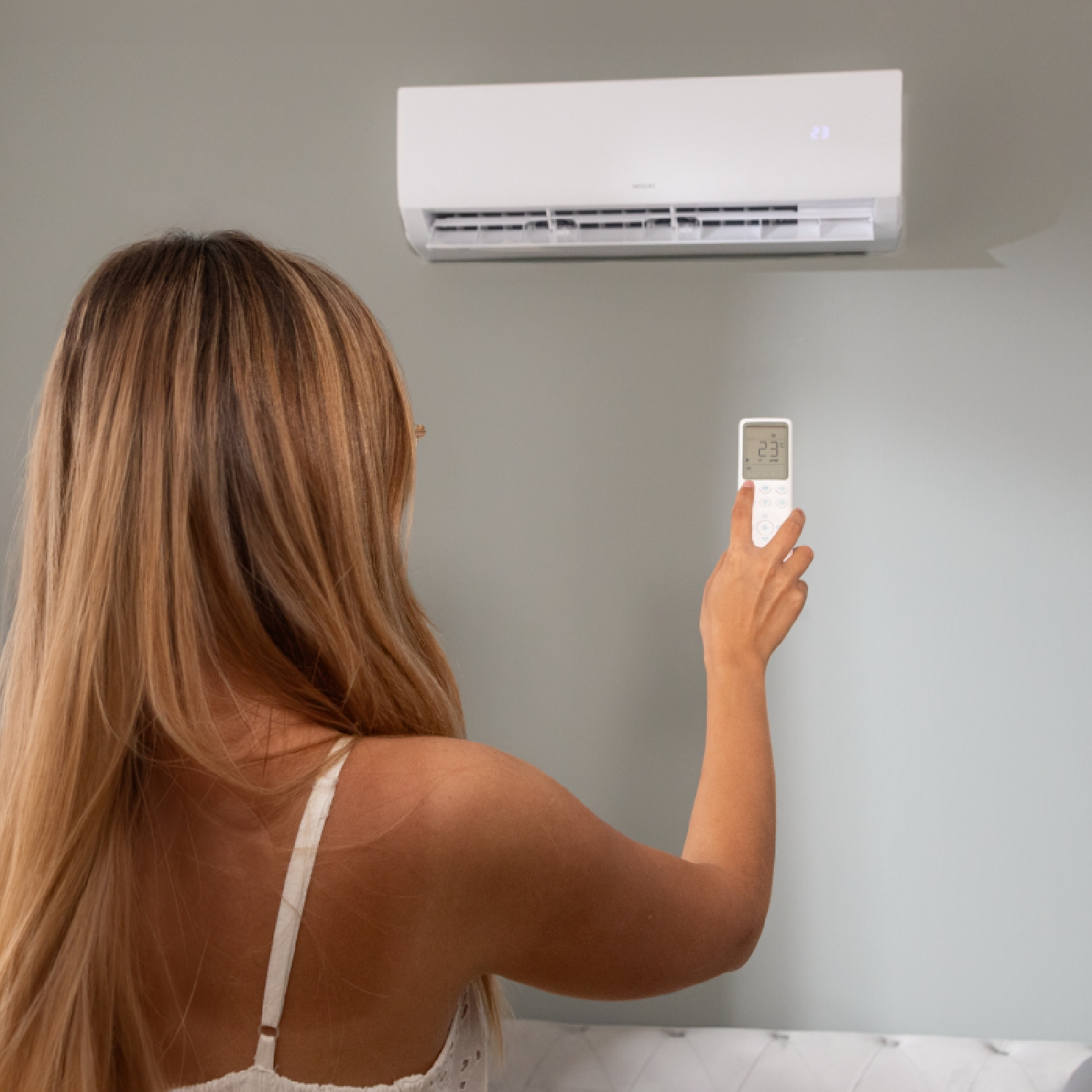 Una mujer ajusta una unidad de aire acondicionado con un mando. La unidad blanca está montada en una pared gris. Sostiene el mando en la mano derecha.