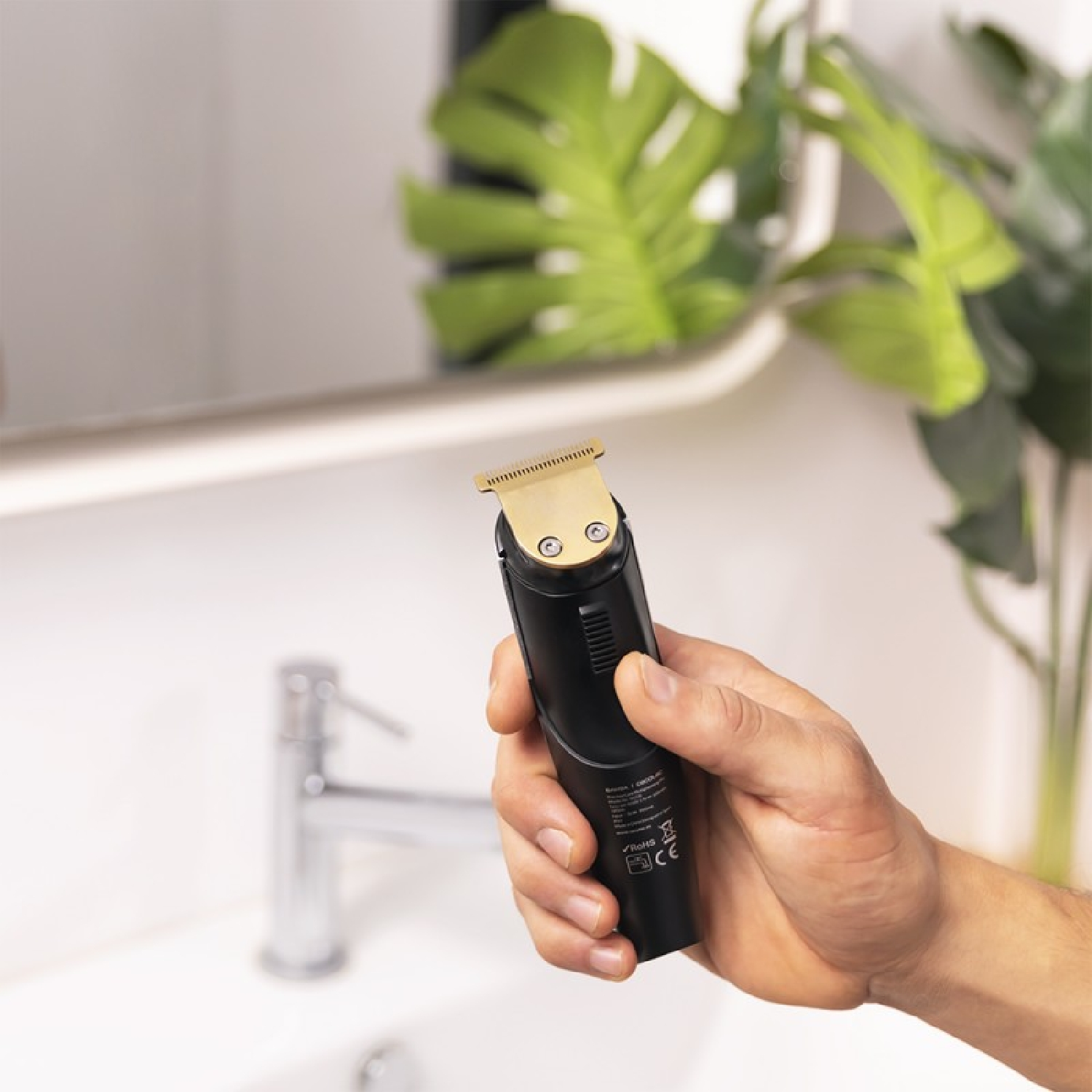 Ręka trzyma czarno-złotą maszynkę do strzyżenia w łazience. W tle lustro, umywalka i zielona roślina.