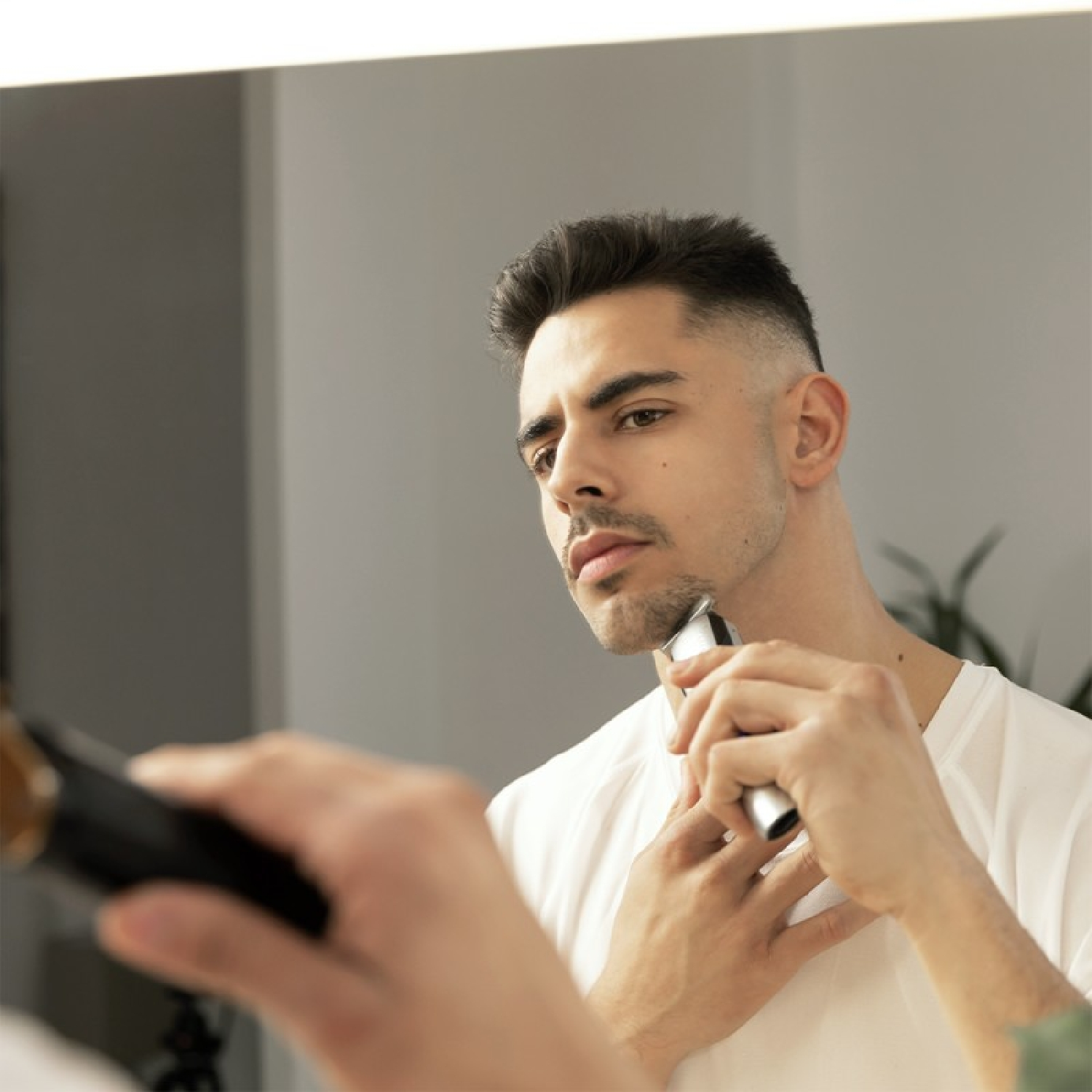 Mężczyzna goli brodę przed lustrem. Trzyma elektryczną maszynkę do golenia przy szyi. Lustro odbija jego twarz.