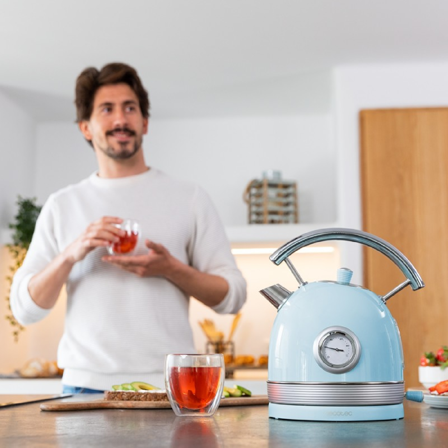 Mężczyzna trzyma szklankę herbaty w kuchni. Niebieski czajnik stoi na blacie. Widoczne jest pieczywo z awokado i herbata w szklance.