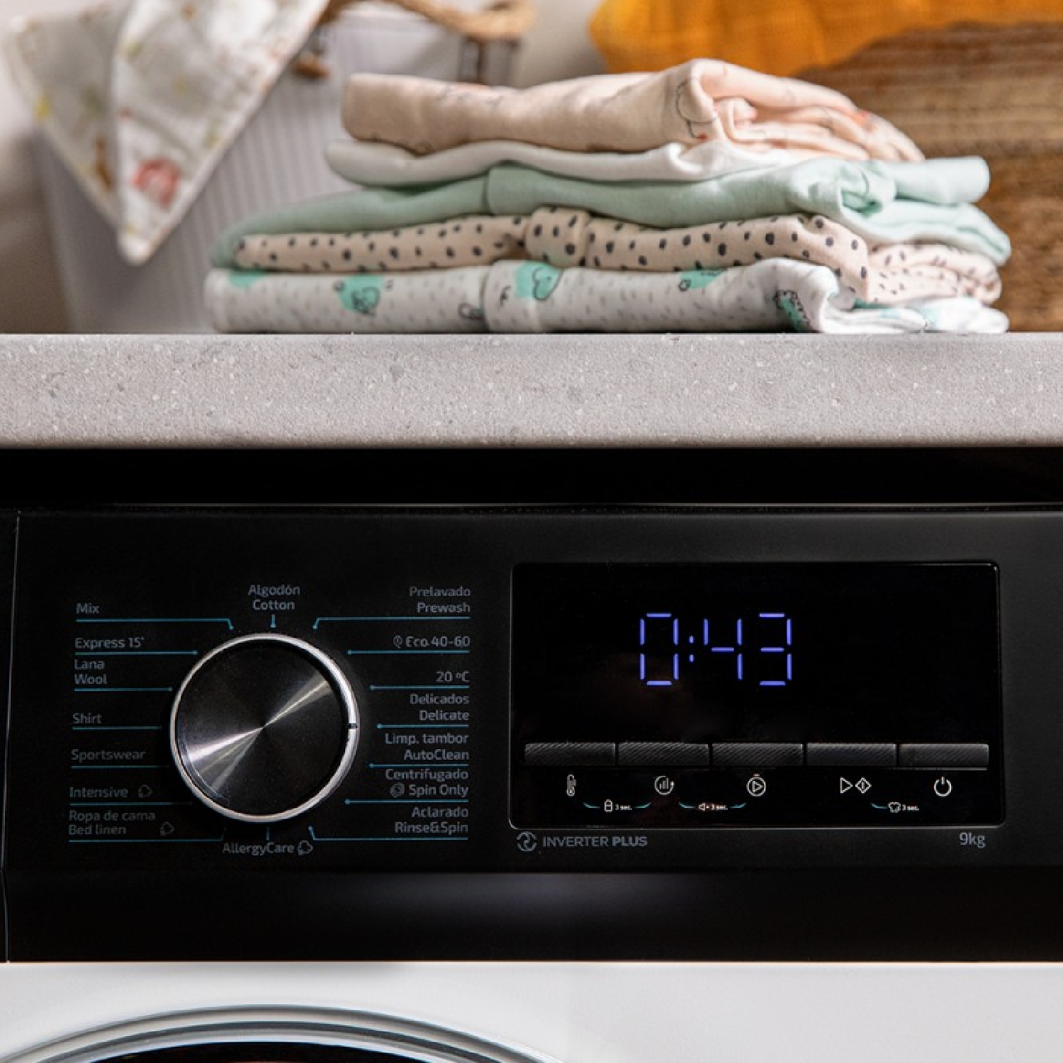 Panel sterowania pralki z wyświetlaczem cyfrowym 0:43. Powyżej stos złożonych ubranek dziecięcych.