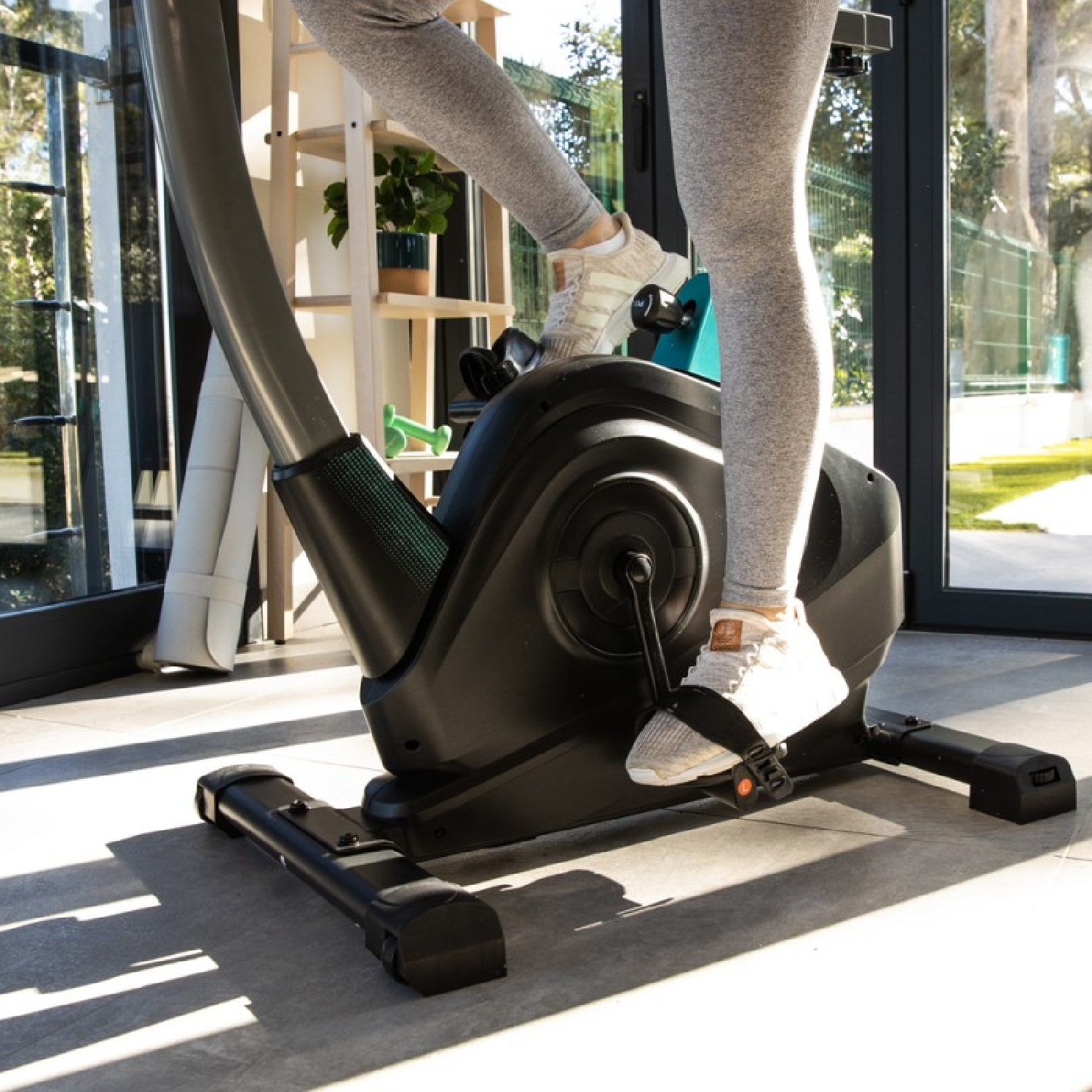 Persona su una cyclette, con leggings grigi e scarpe da ginnastica bianche. La bici è nera. Lo sfondo mostra una stanza con piante.