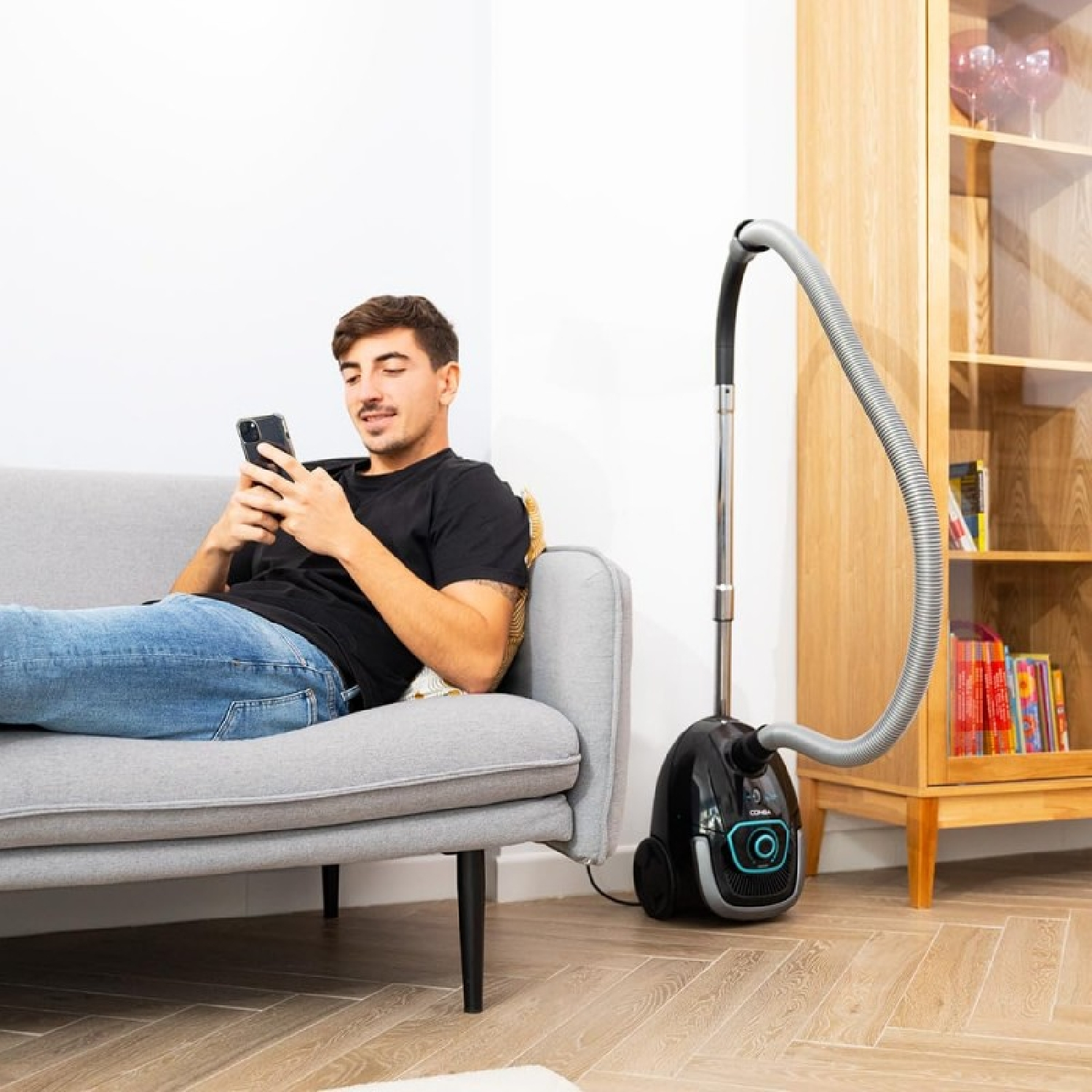 Een man zit op een bank en kijkt naar zijn telefoon. Een stofzuiger staat in de buurt en een houten kast op de achtergrond.