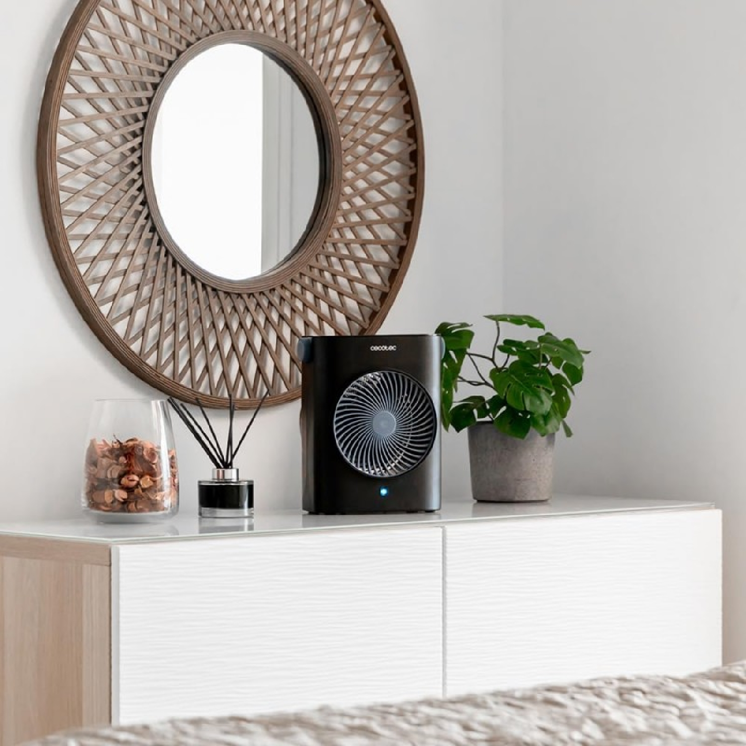 Scena sypialni z okrągłym lustrem, czarnym wentylatorem, rośliną i dyfuzorem na białej komodzie. Lustro ma plecioną ramę.