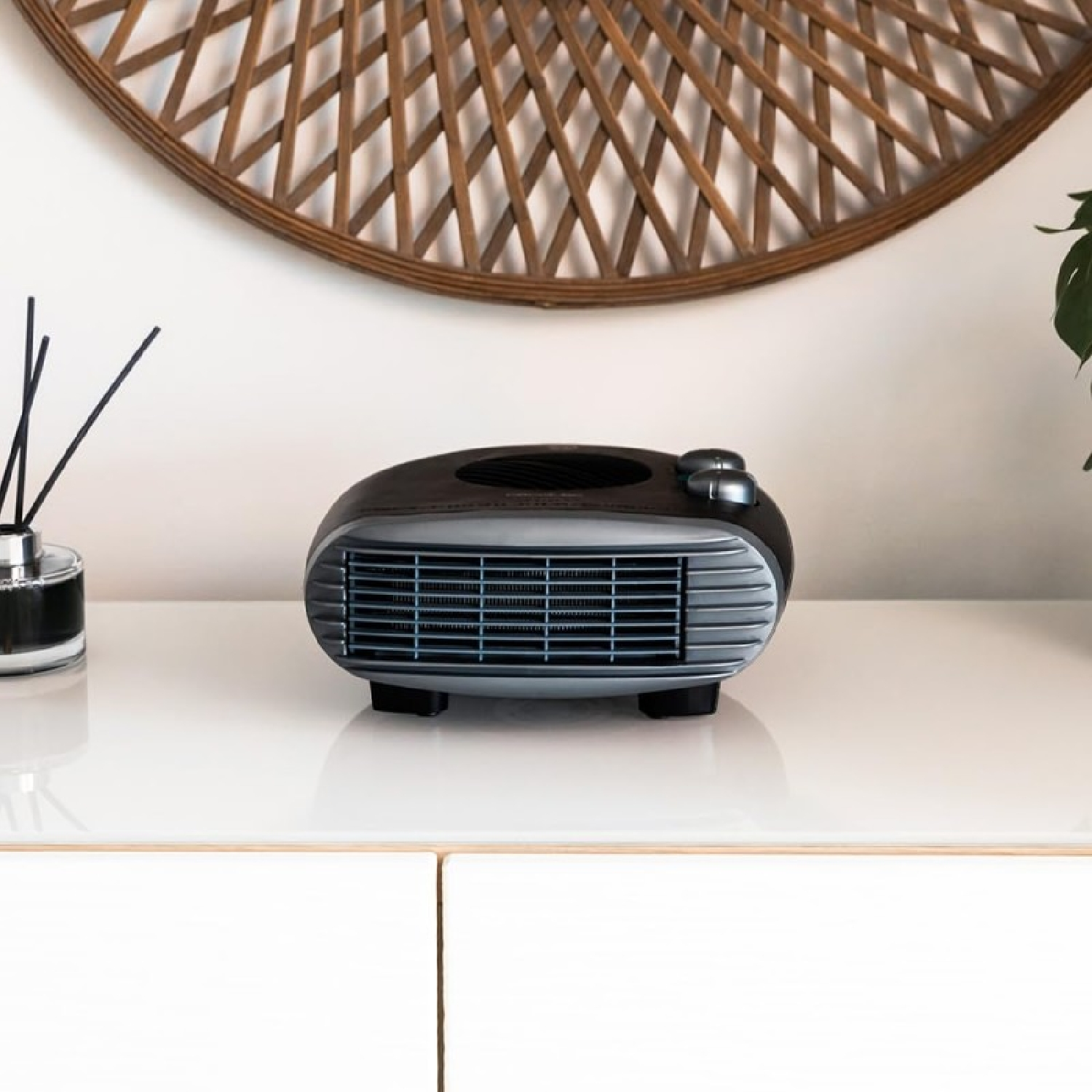 Czarny i srebrny grzejnik stoi na białej powierzchni. Za nim znajduje się tkana okrągła dekoracja ścienna. Po lewej stronie znajduje się dyfuzor trzcinowy.