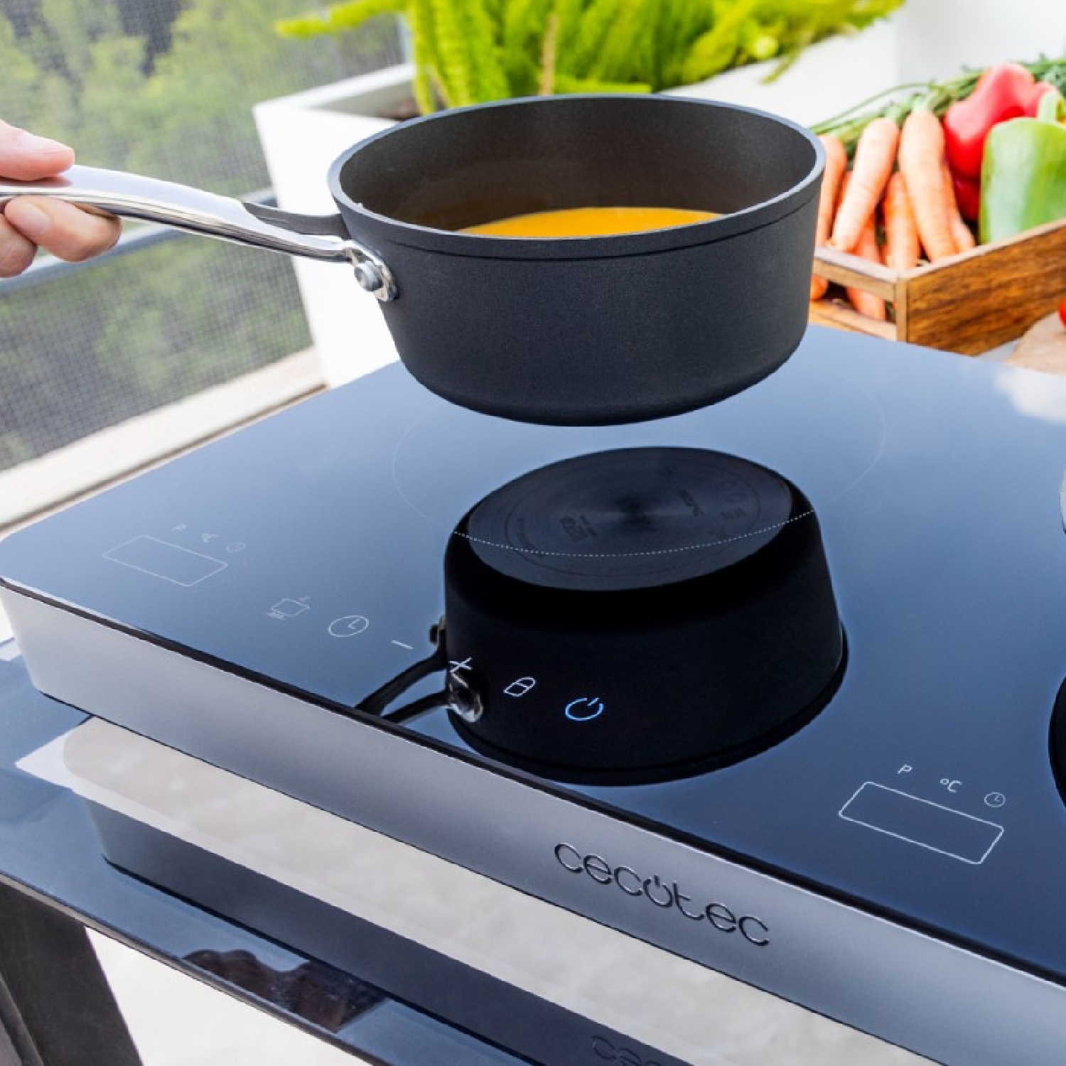 Ręka trzyma czarny garnek z pomarańczowym płynem nad czarną płytą indukcyjną. Warzywa w drewnianej skrzyni w tle.