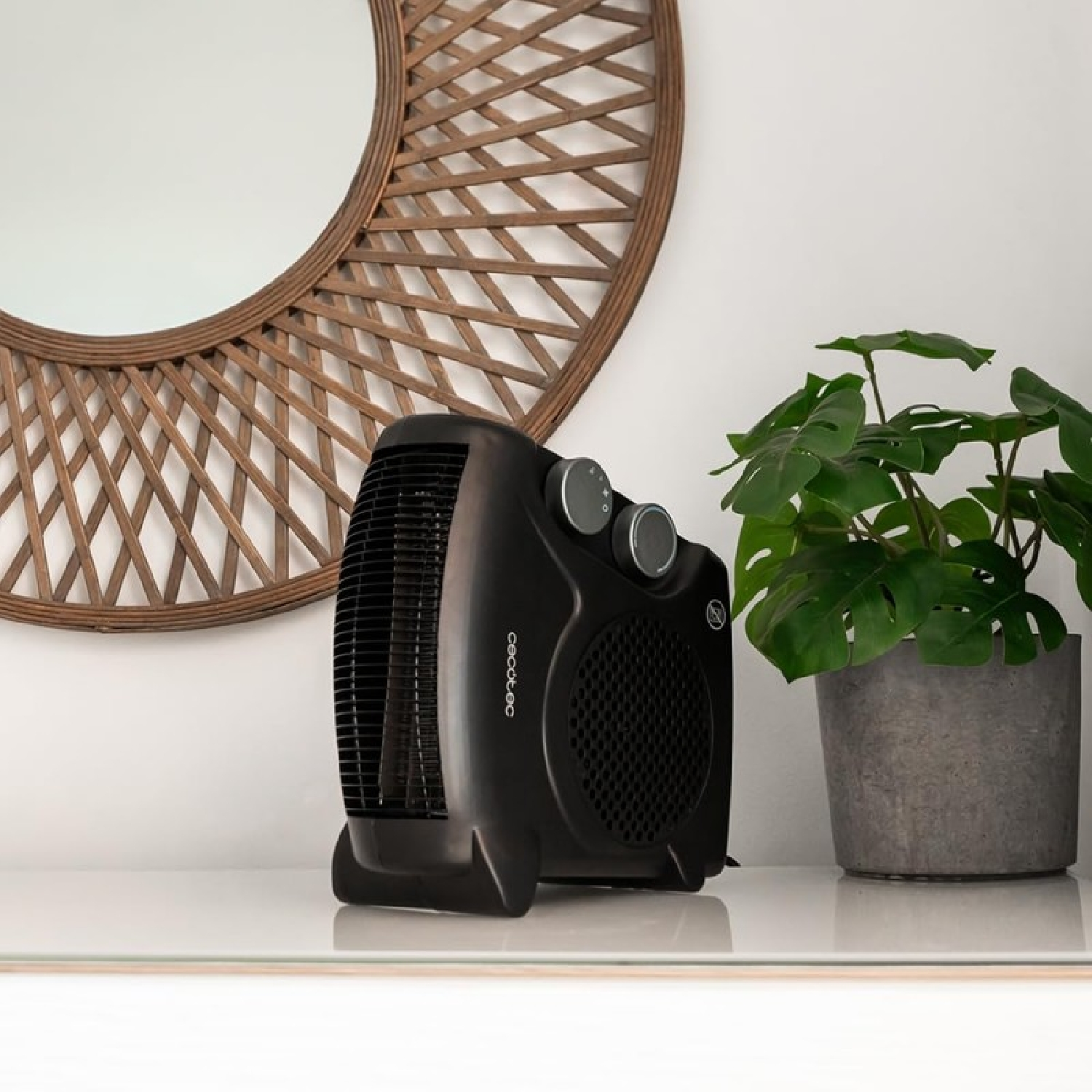 Un termoventilatore nero è su un ripiano bianco vicino a una pianta in vaso. Uno specchio rotondo intrecciato è appeso sopra.
