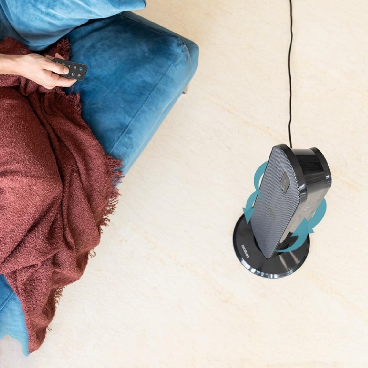Una persona sostiene un mando a distancia. Un ventilador está en el suelo, junto a un sofá azul y una manta roja.