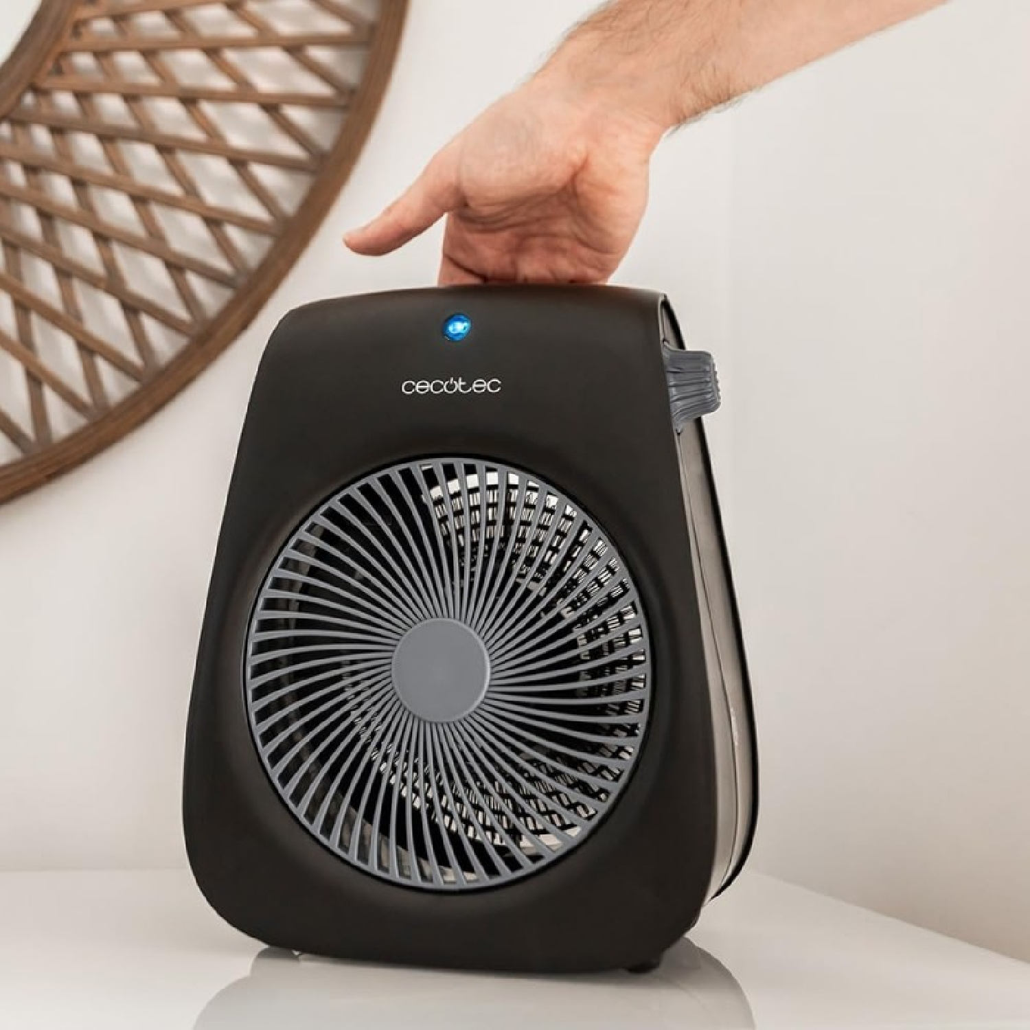 Ręka dotyka czarnego wentylatora z szarą kratką. Po lewej stronie znajduje się drewniana, dekoracyjna dekoracja ścienna.