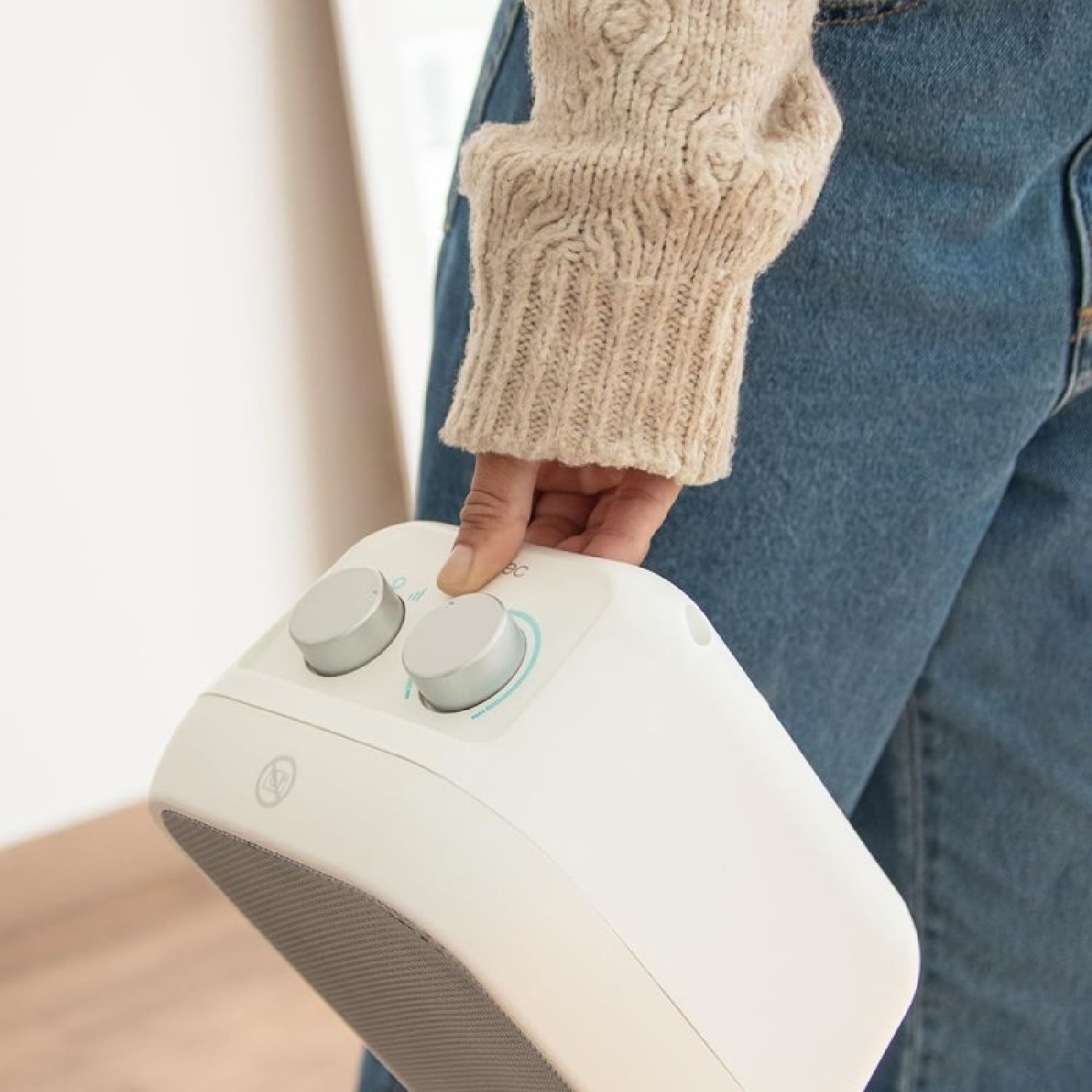 Osoba trzyma biały przenośny grzejnik. Osoba ma na sobie beżowy sweter i niebieskie dżinsy. Grzejnik ma dwa pokrętła i szarą kratkę.