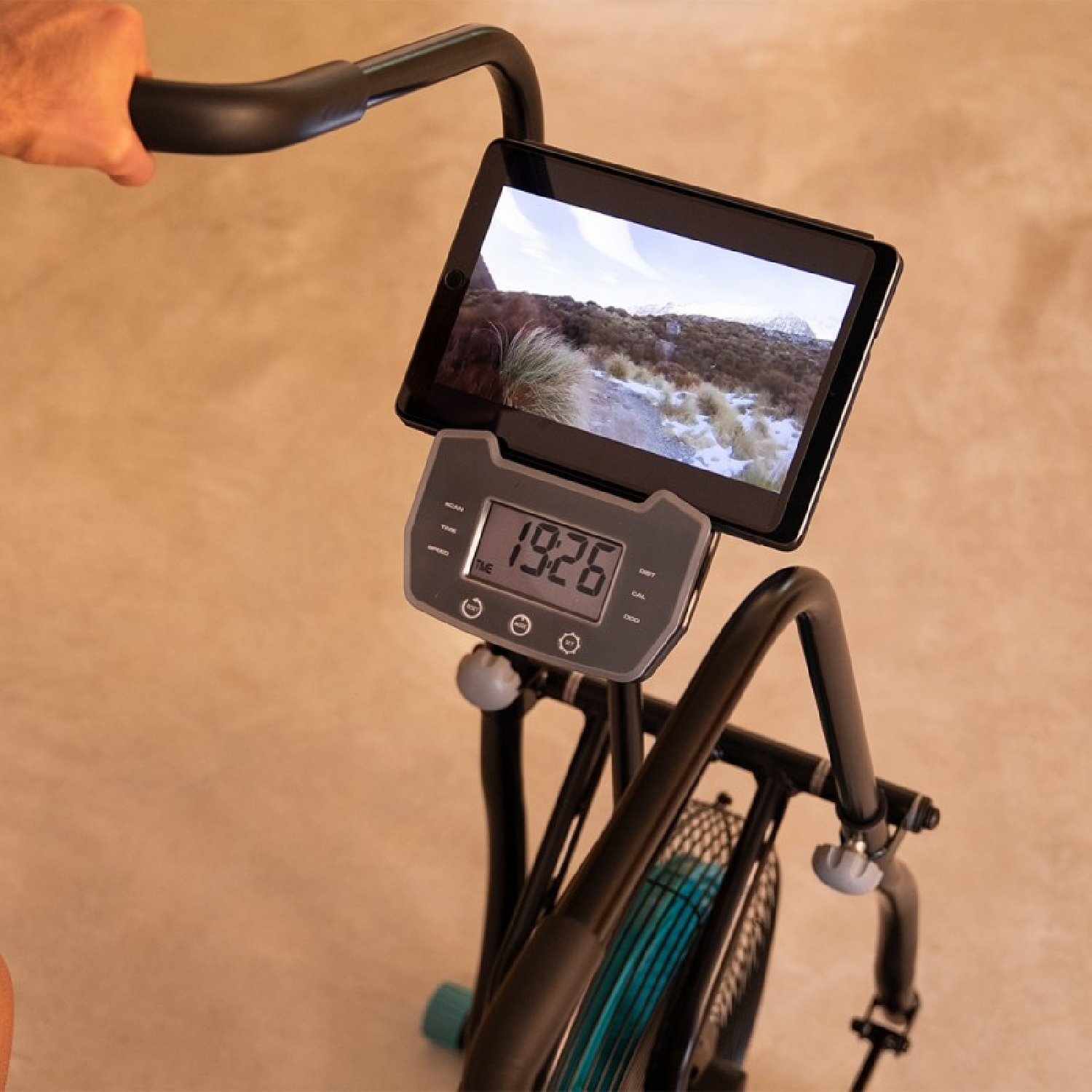 Bicicletta da camera con tablet che mostra un paesaggio. Mano sul manubrio. Il display mostra l'ora. Accenti neri e turchesi sulla bici.