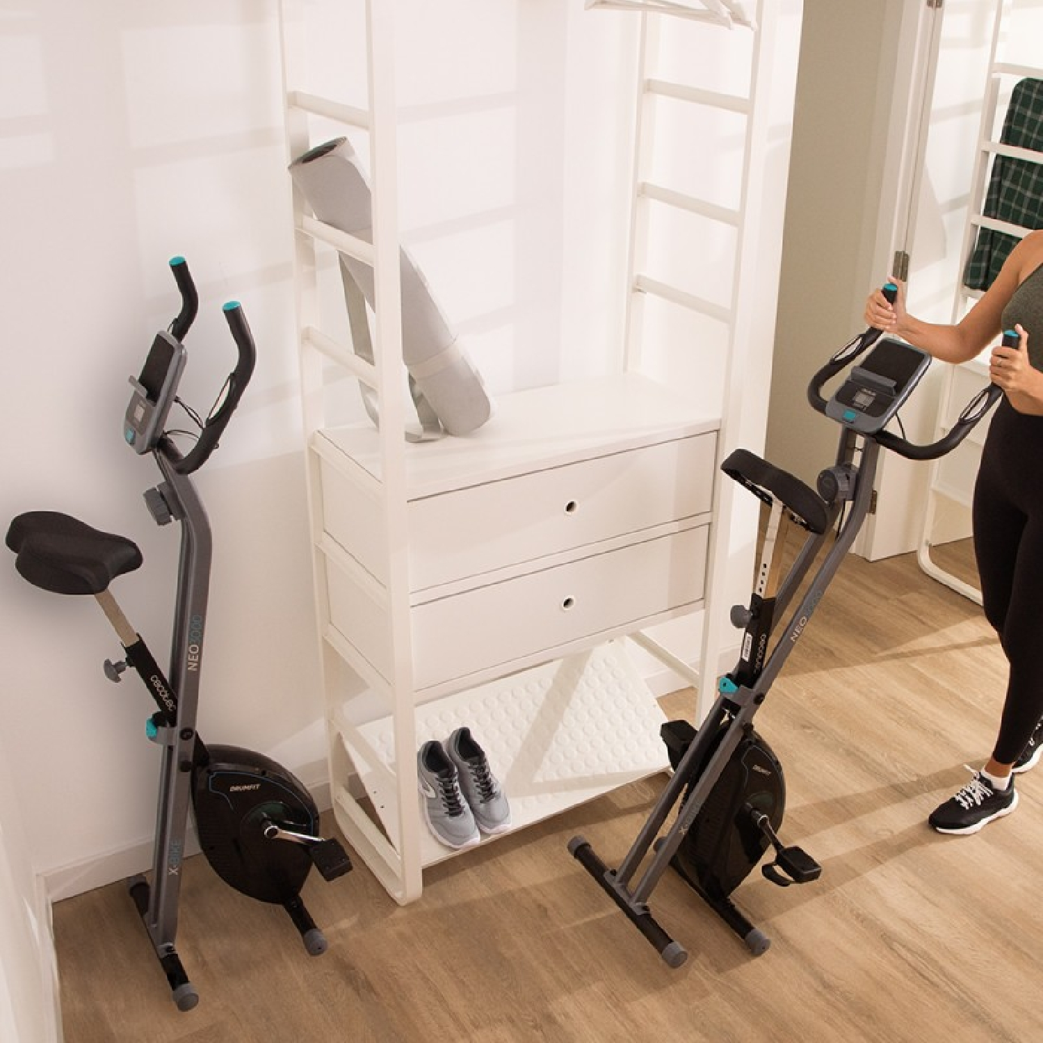 Dwa rowery stacjonarne w pokoju. Kobieta stoi obok jednego. Szafa ze złożoną matą, butami i wieszakiem w tle.