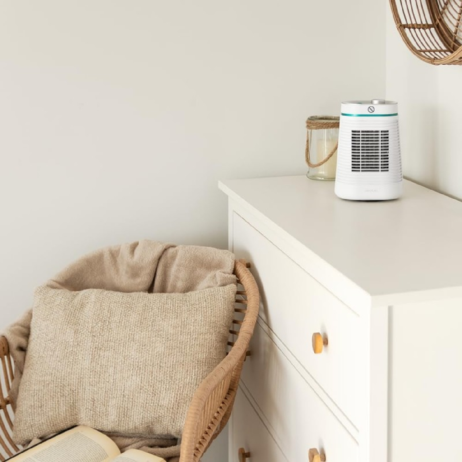 Un termoventilatore bianco su un comò bianco. A sinistra, una sedia in vimini con cuscino e coperta. Una candela e decorazioni murali visibili.