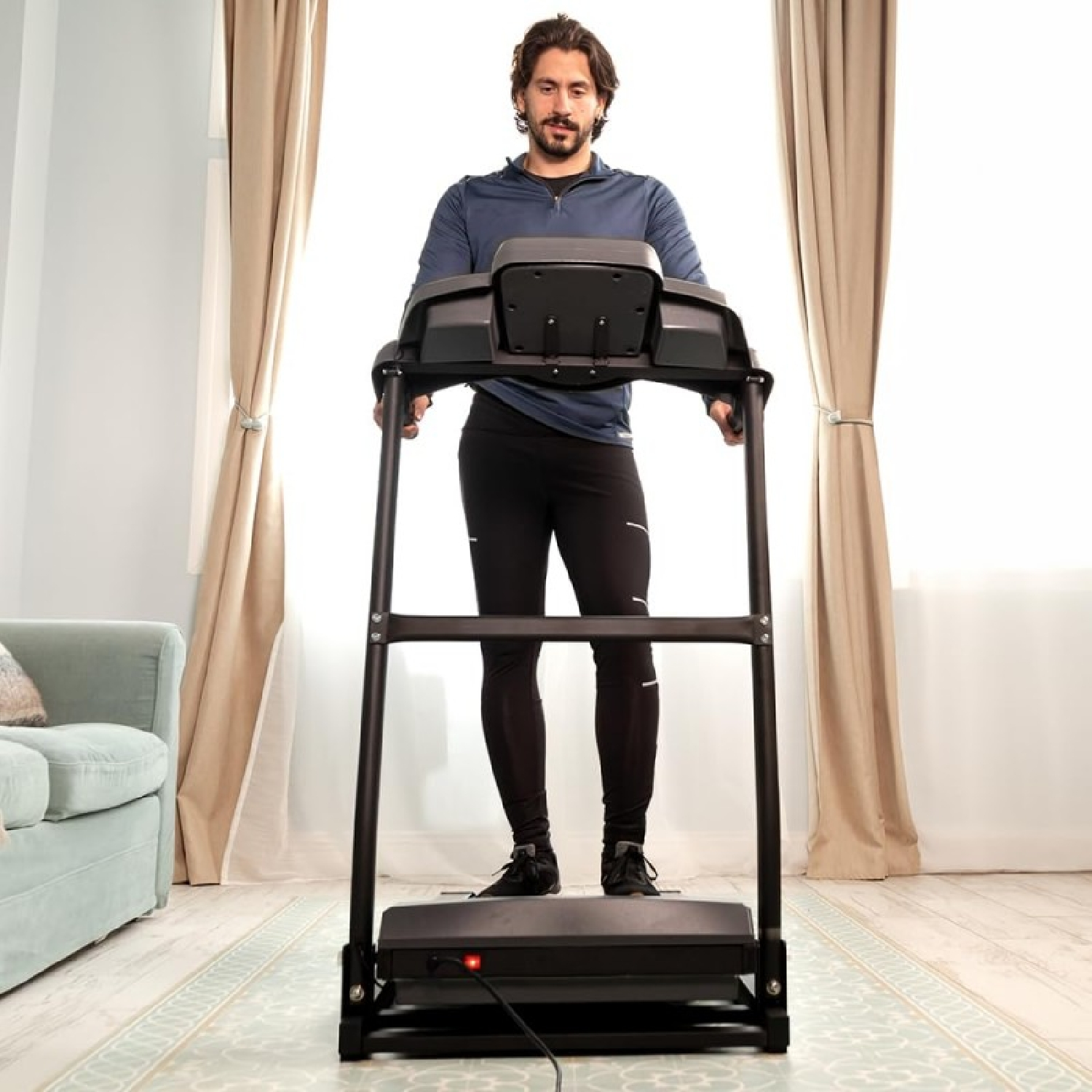 Mężczyzna stoi na bieżni w pomieszczeniu. Ma na sobie czarne spodnie sportowe i niebieską koszulę. Za nim są beżowe zasłony.
