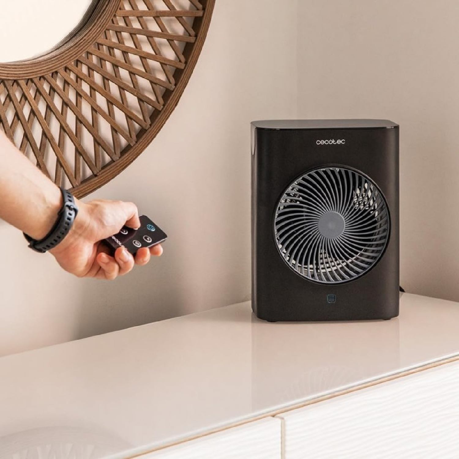 Una mano con un telecomando controlla un ventilatore nero. Uno specchio decorativo è sullo sfondo. Il ventilatore è su una superficie bianca.
