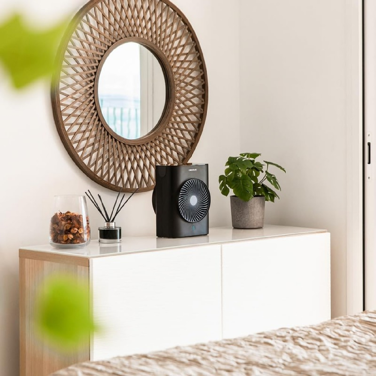 Scena di camera da letto con specchio rotondo, ventilatore nero, pianta e oggetti decorativi su un mobile bianco. Il letto in primo piano.