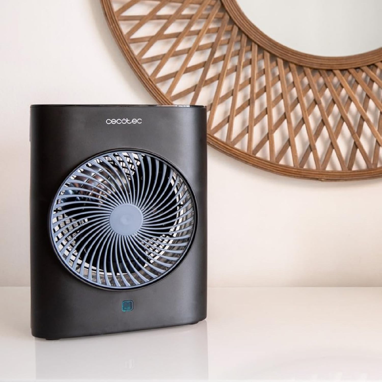 Un ventilatore nero con una griglia blu è su una superficie bianca. Uno specchio con cornice in legno è appeso sullo sfondo.