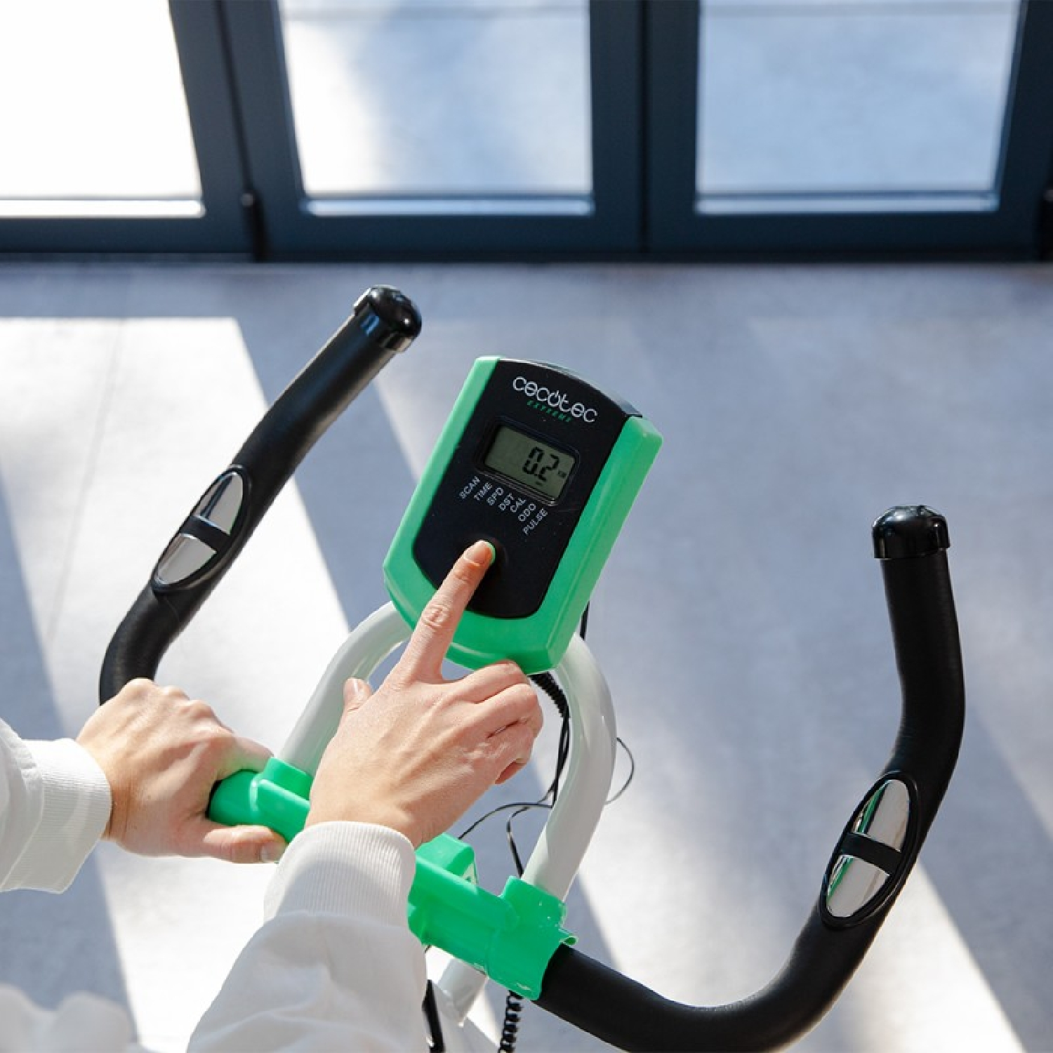 Le mani di una persona sono sul manubrio di una cyclette. Il display indica 0,2. La bici è verde e bianca.