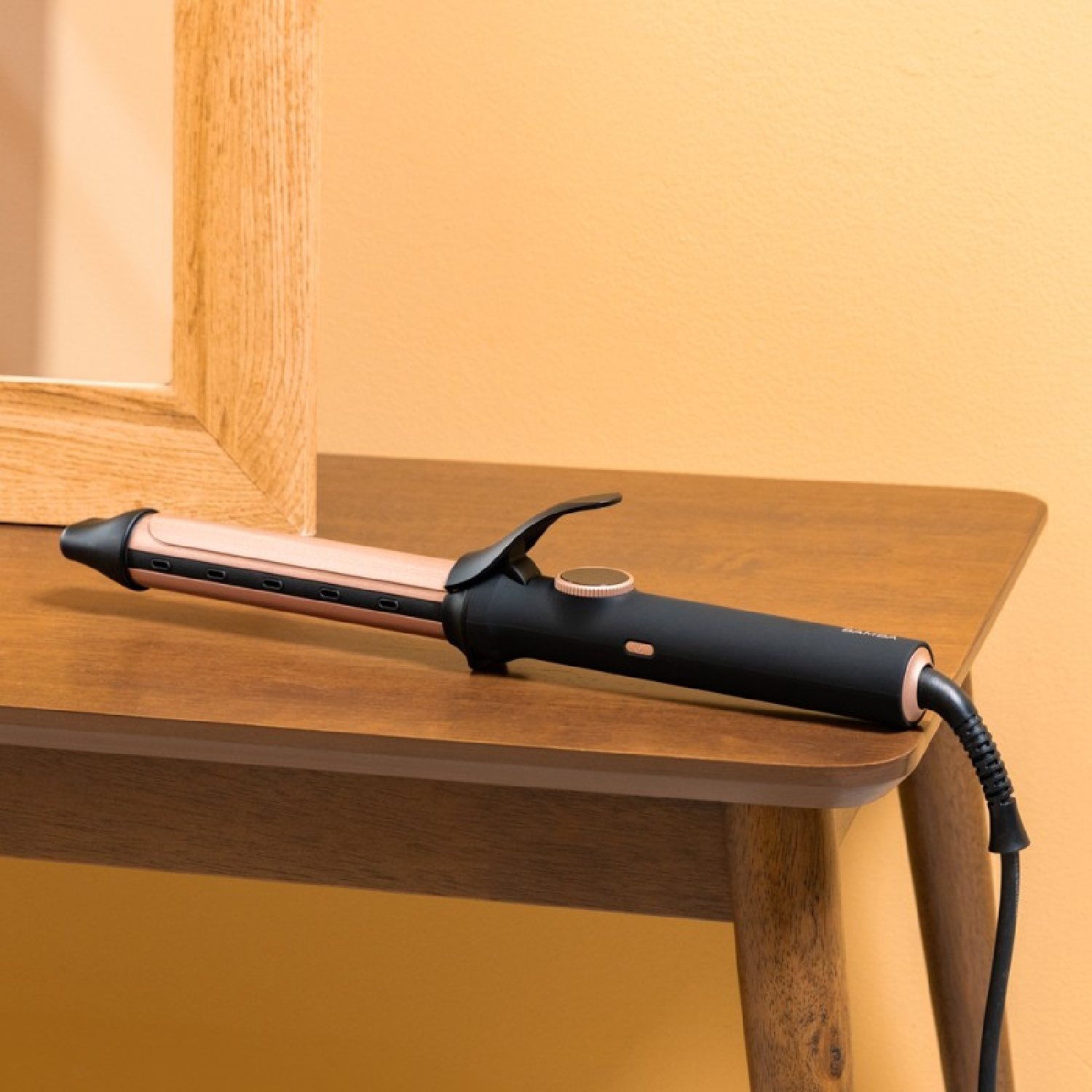 Una plancha para rizar el cabello está sobre una mesa de madera marrón. Un espejo con marco de madera está a la izquierda de la mesa. La pared es amarilla.