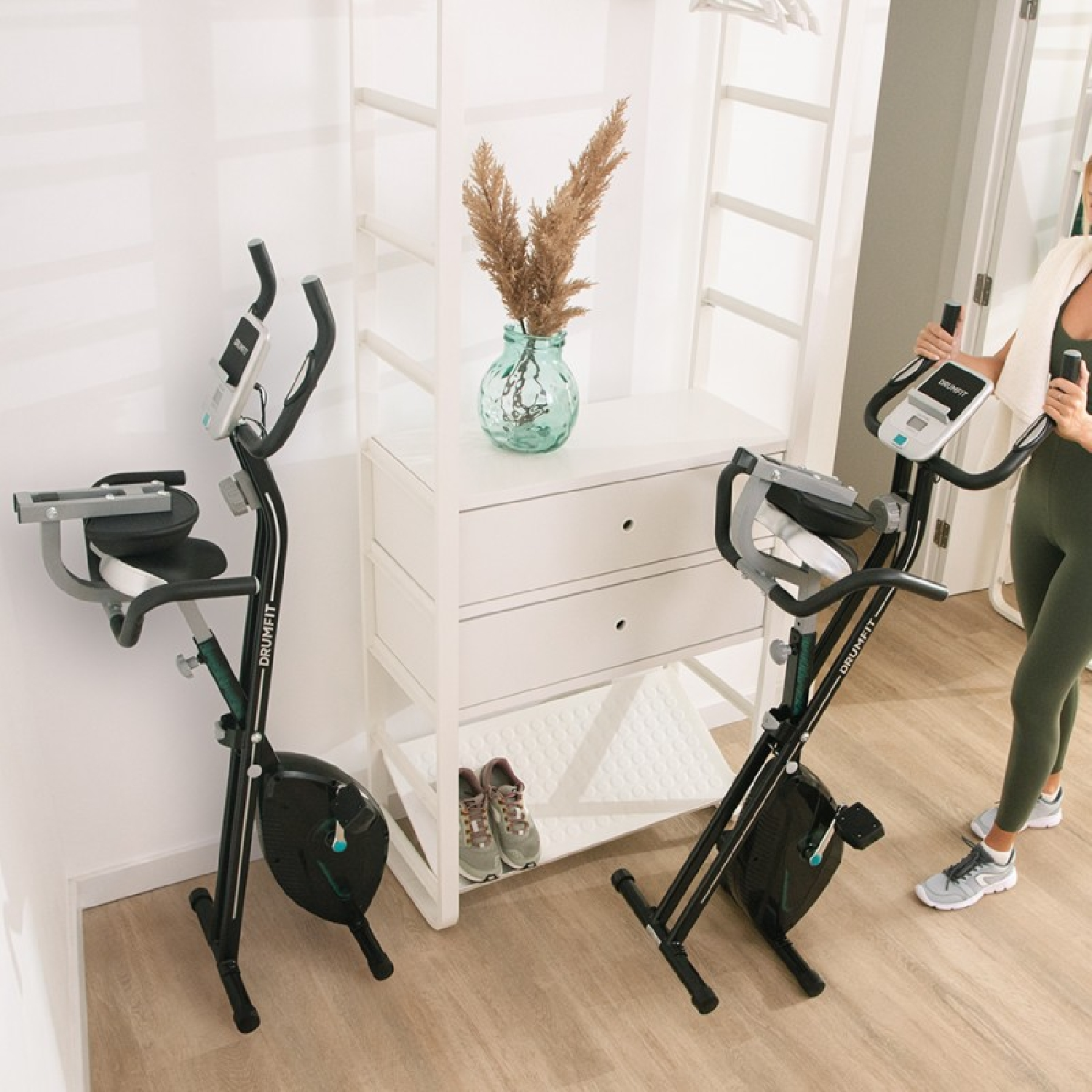 Dos bicicletas de ejercicio negras en una habitación. Una mujer está al lado. Una estantería blanca con un jarrón y zapatos a un lado.