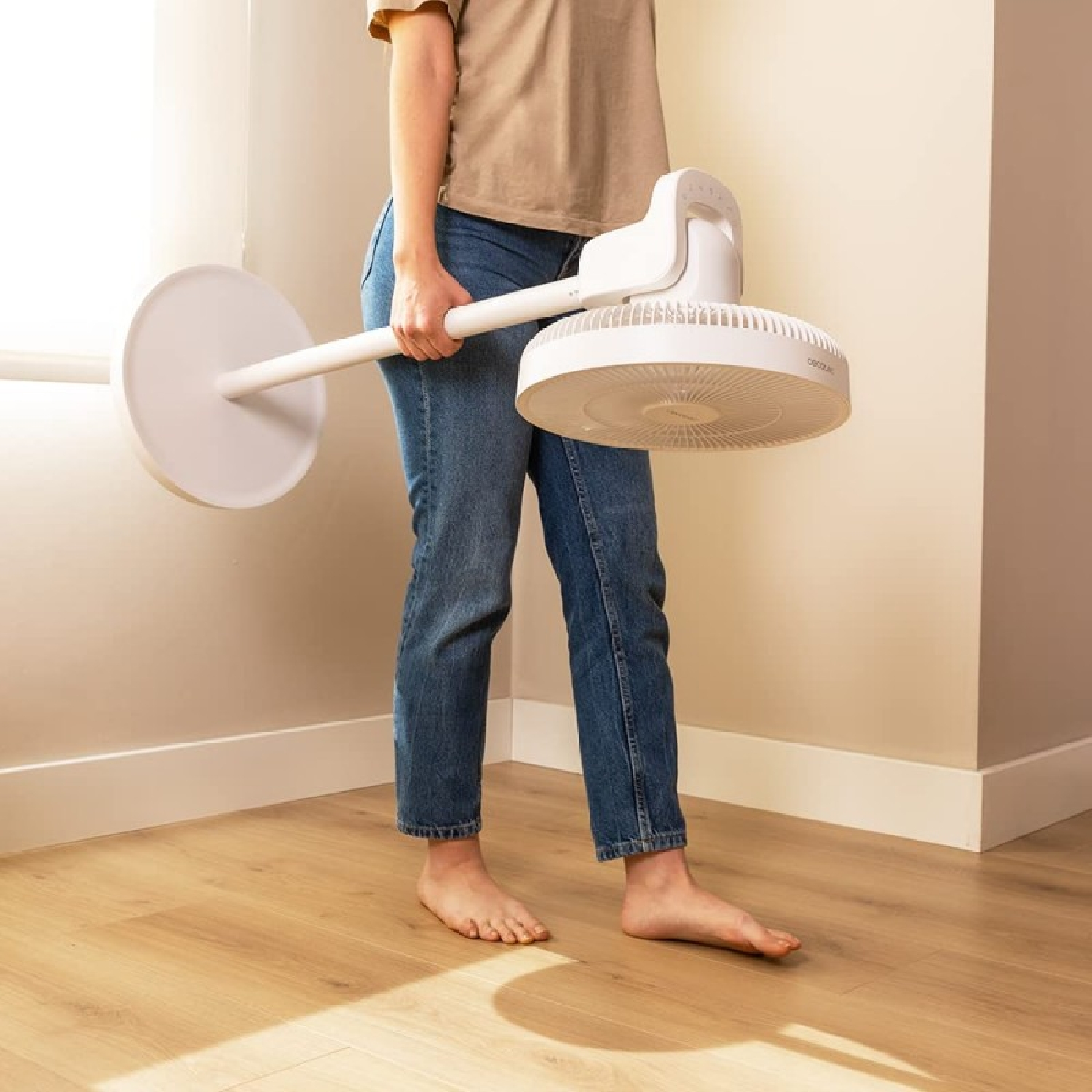 Osoba trzyma biały wentylator. Stoi boso na drewnianej podłodze, blisko okna. Wentylator jest duży i okrągły.