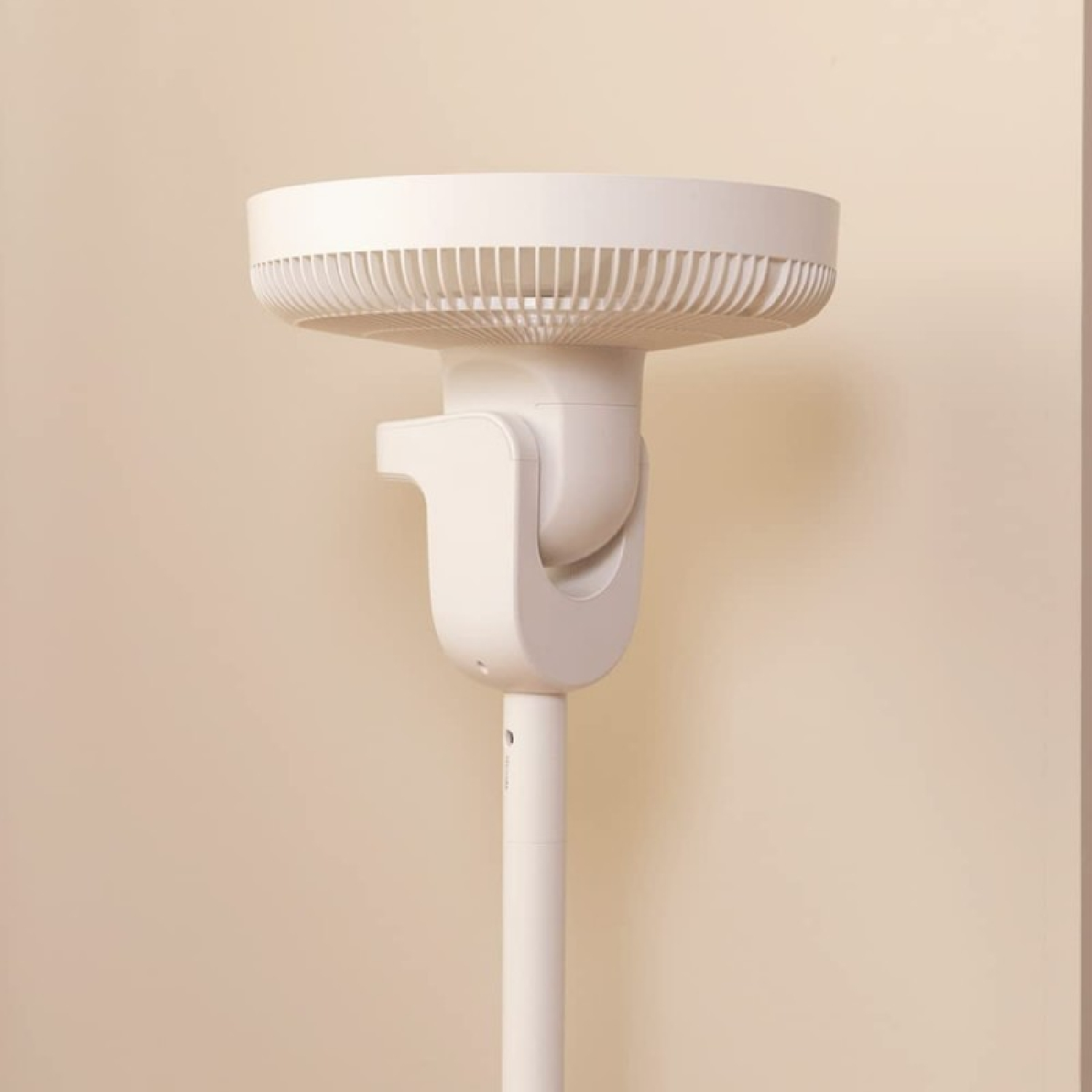Biały wentylator stojący stoi przed beżową ścianą. Wentylator ma okrągłą głowicę z otworami wentylacyjnymi i zakrzywioną podstawę.