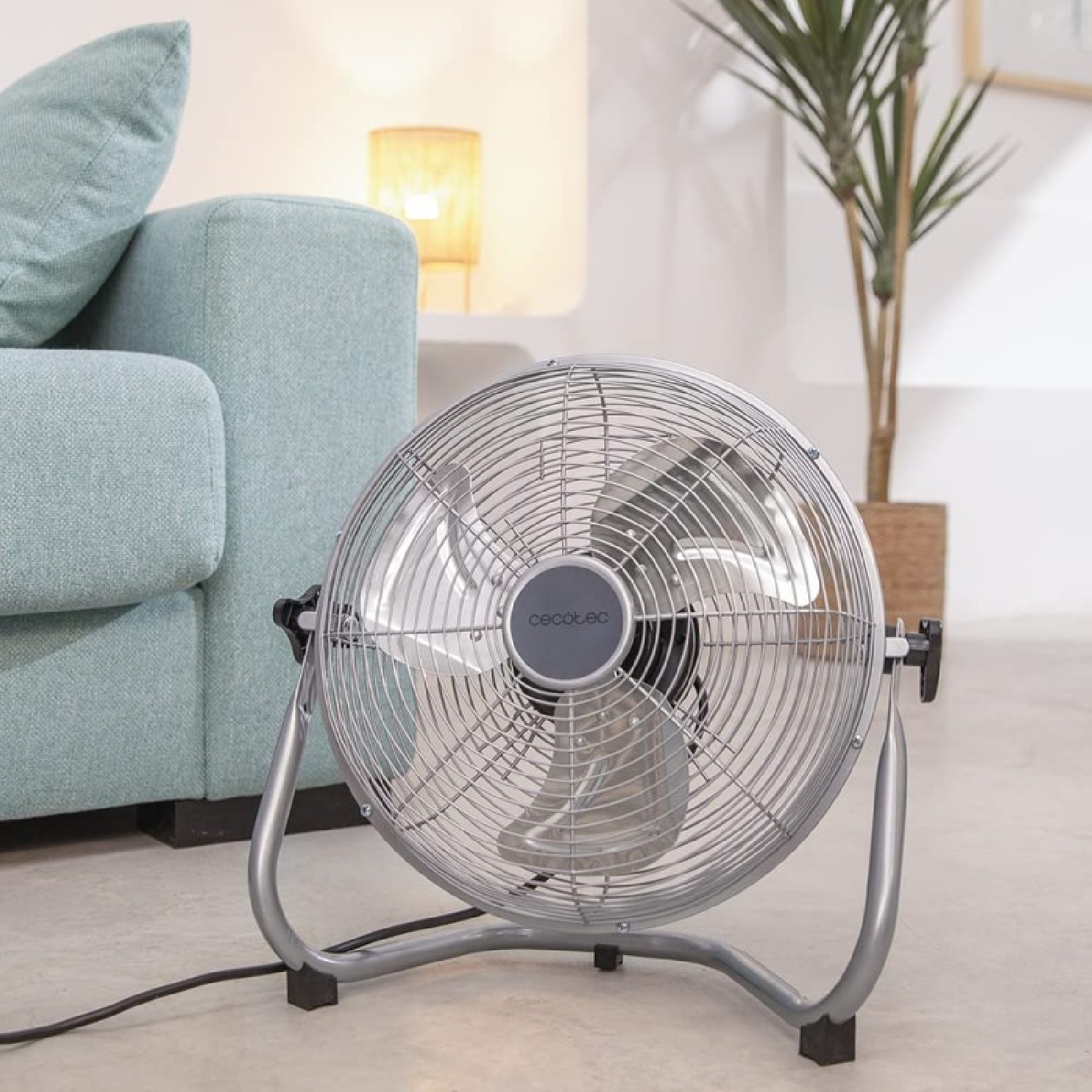 Srebrny wentylator stoi na podłodze, przed turkusową sofą i rośliną w brązowej doniczce. Z tyłu świeci lampa.