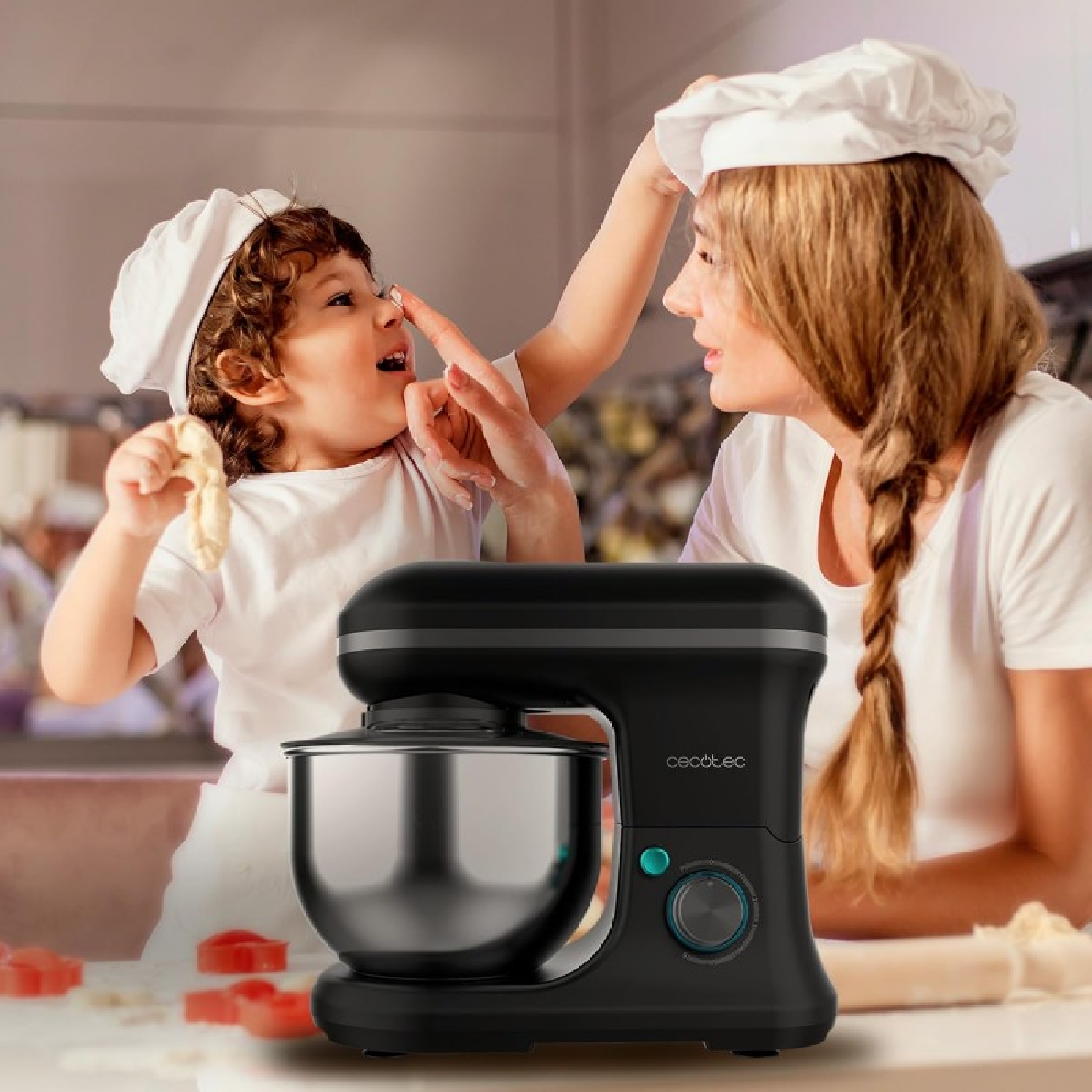 Una donna e un bambino con cappelli da chef interagiscono mentre cucinano. Un mixer nero è in primo piano. Tagliapasta a forma di cuore rossi sul bancone.