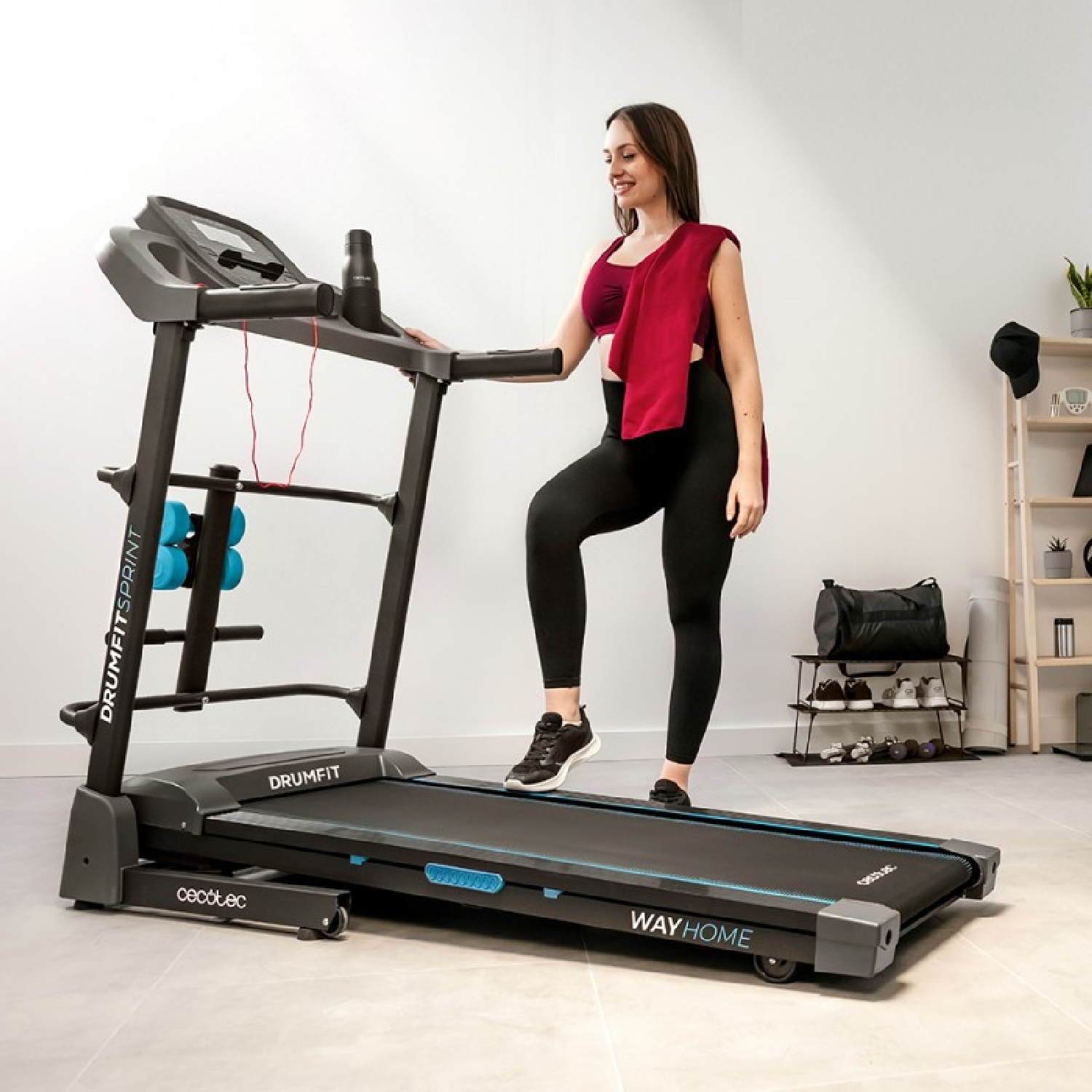 Kobieta na bieżni, czerwony ręcznik na ramieniu. Czarne legginsy, stanik sportowy. Pokój z butami, torbą, półkami i butelką wody.