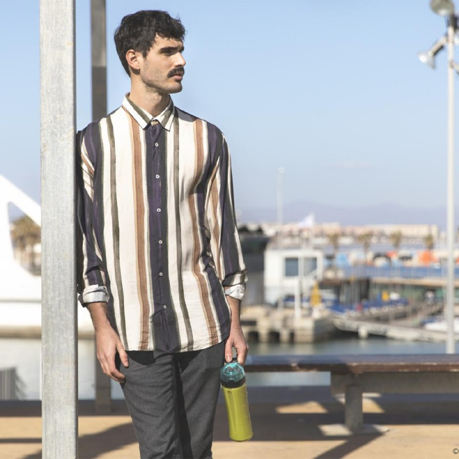 Un hombre con camisa a rayas se apoya en un poste, sosteniendo una botella de agua. Fondo: un puerto deportivo con barcos bajo un cielo azul.