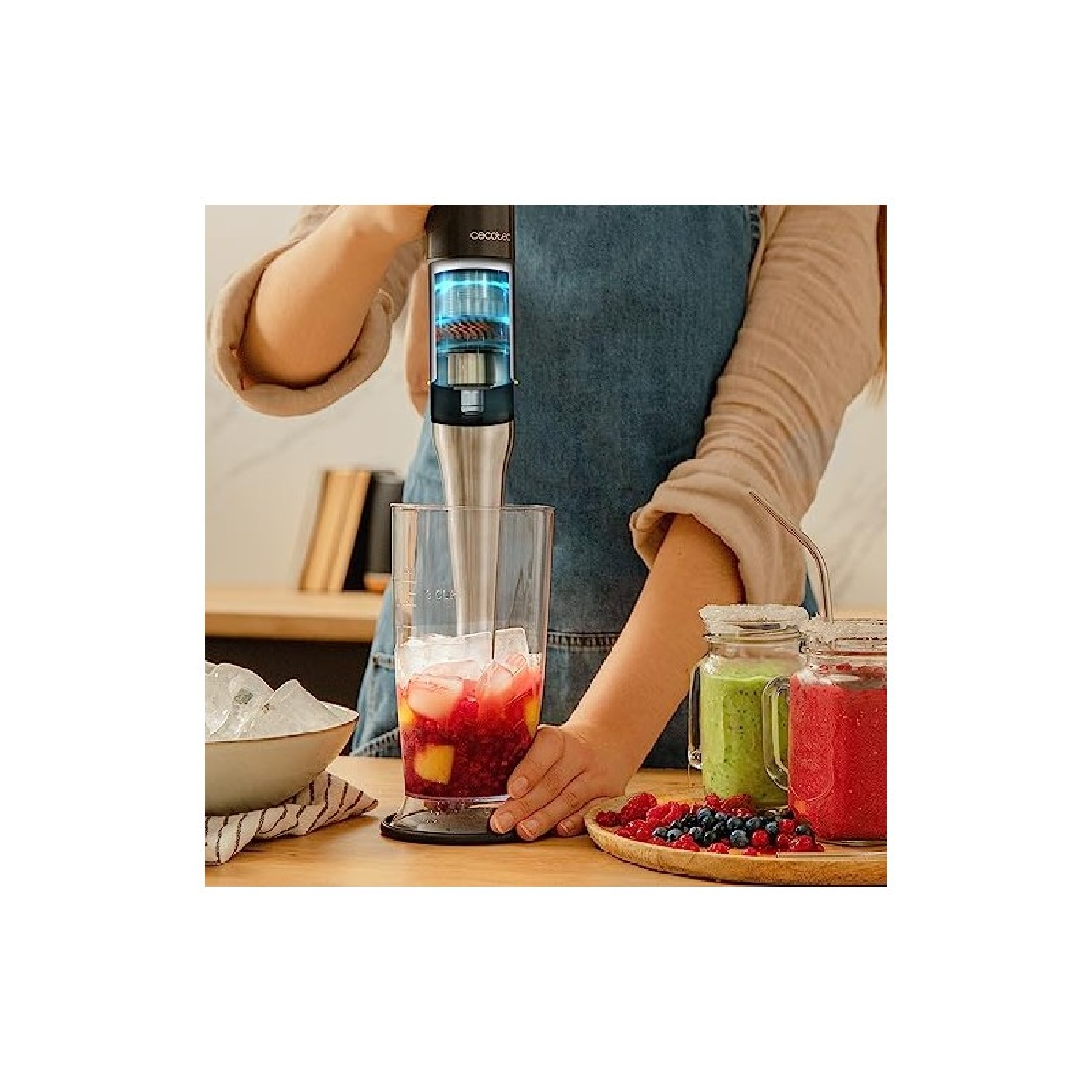 Eine Person mixt mit einem Mixer ein Getränk mit Eis und Früchten in einem Glas. Gläser mit Smoothies und eine Schale mit Eis stehen auf dem Tisch.