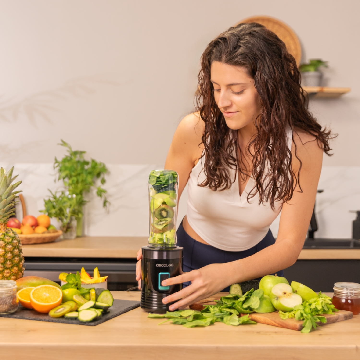Donna che prepara un frullato con un frullatore. Frutta e verdura sul tavolo. Ananas, arancia, kiwi e mele sono visibili.
